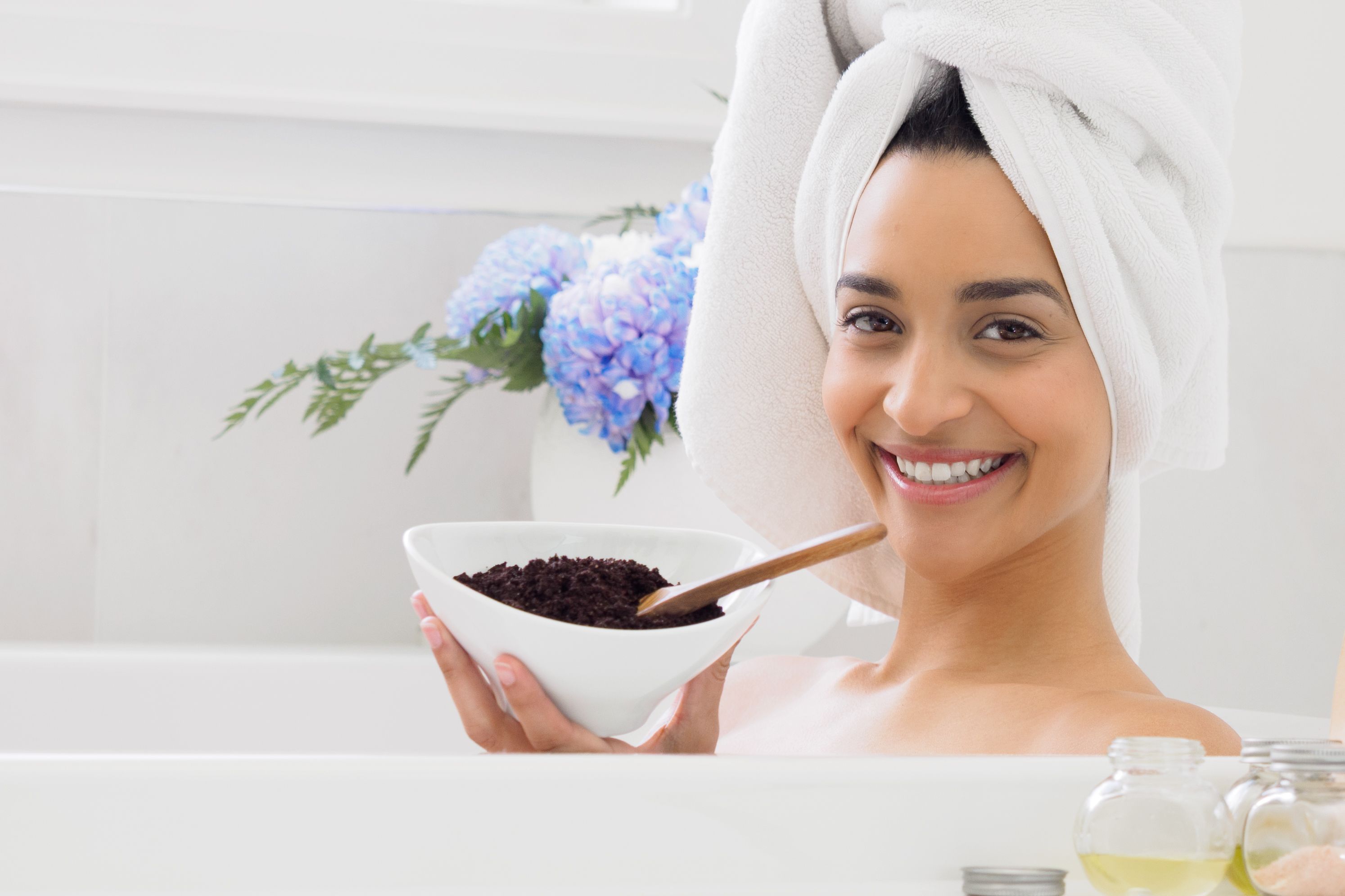 Smiling woman in bathtub with towel turban holding coffee scrub bowl, enjoying a spa day.