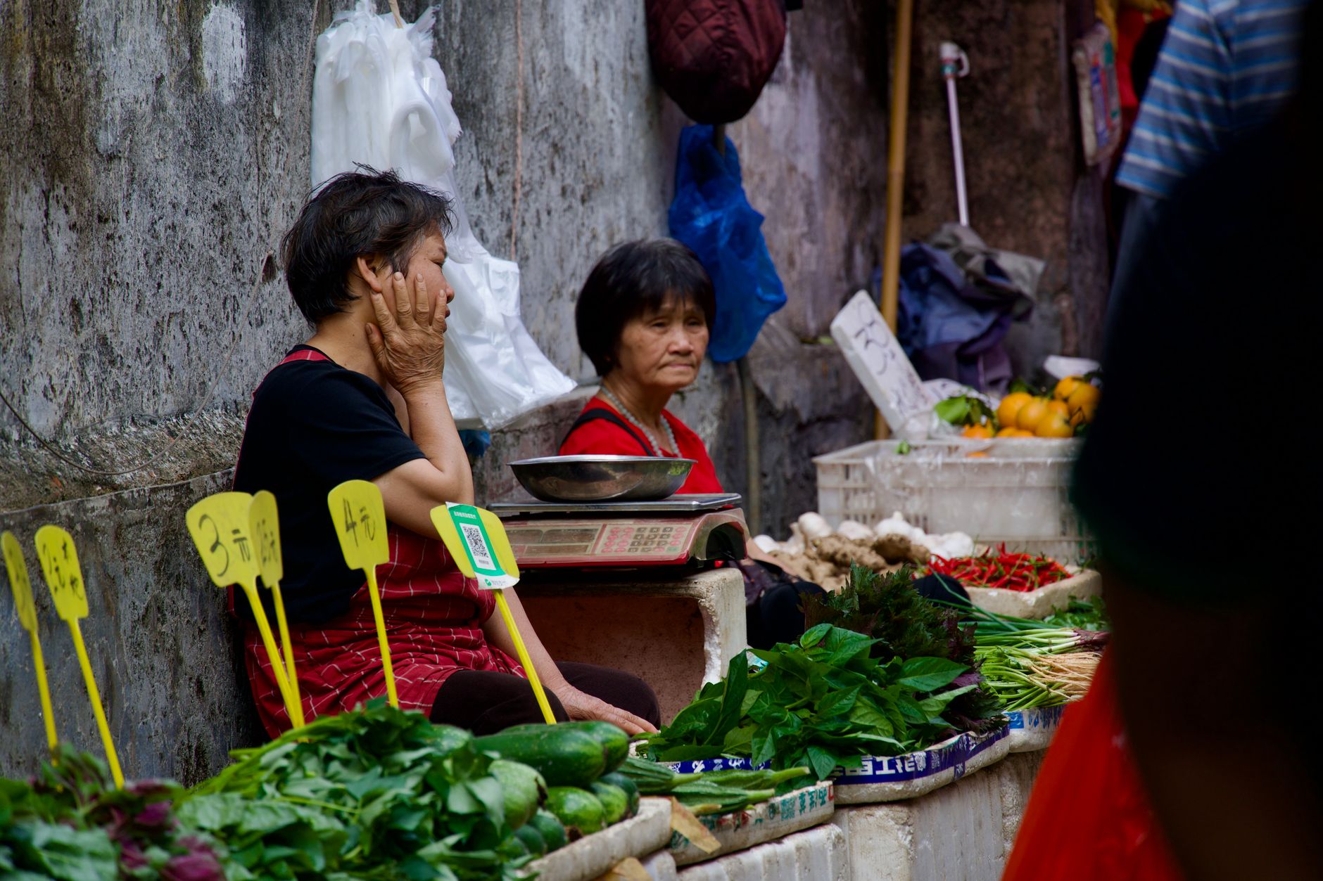Two women at an outdoor market stall with fresh produce and signs.