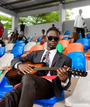 A stylish man in sunglasses plays guitar in a vibrant stadium with colorful seats.