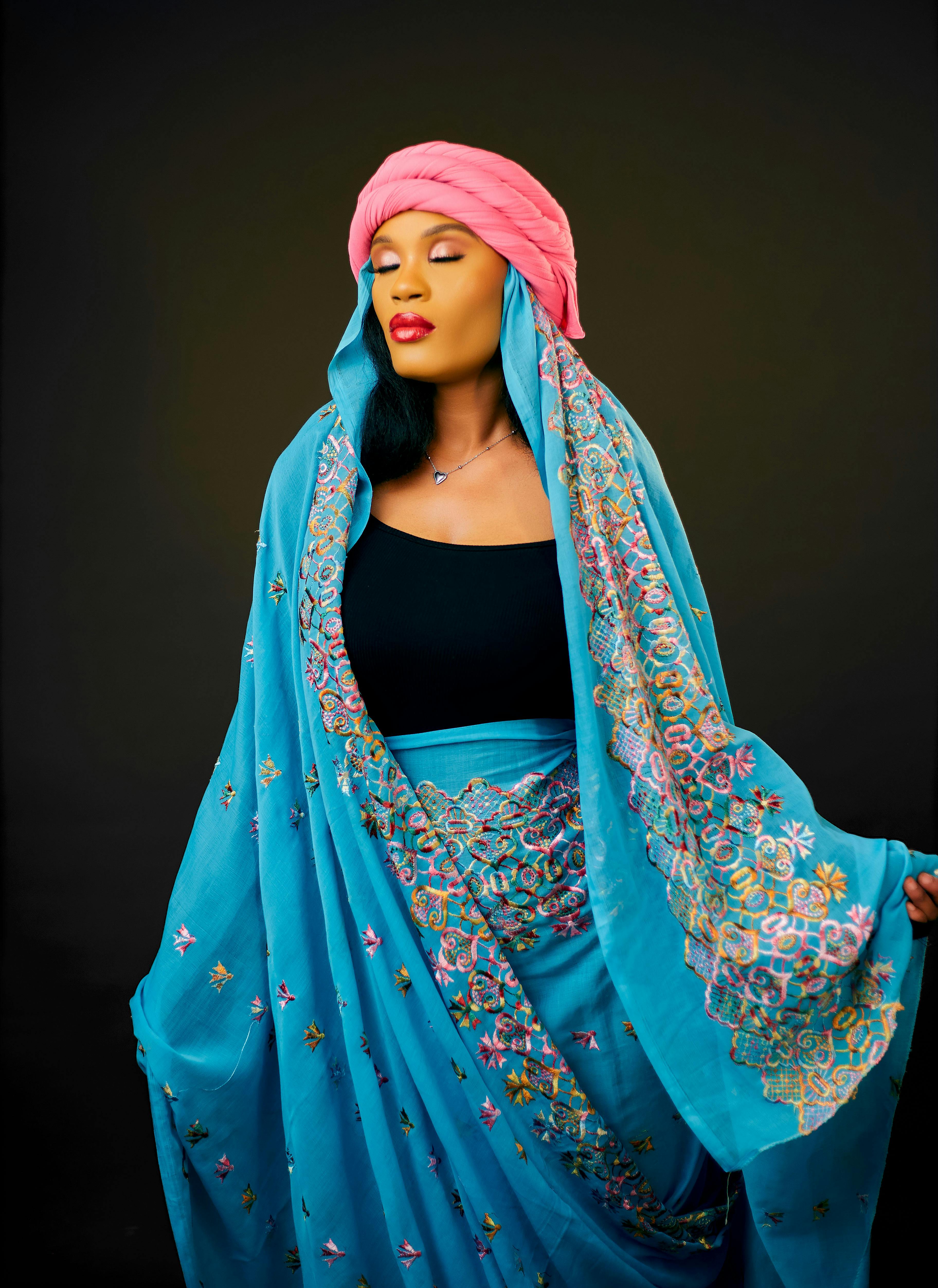 Woman in blue floral embroidered shawl with pink headdress on dark background.