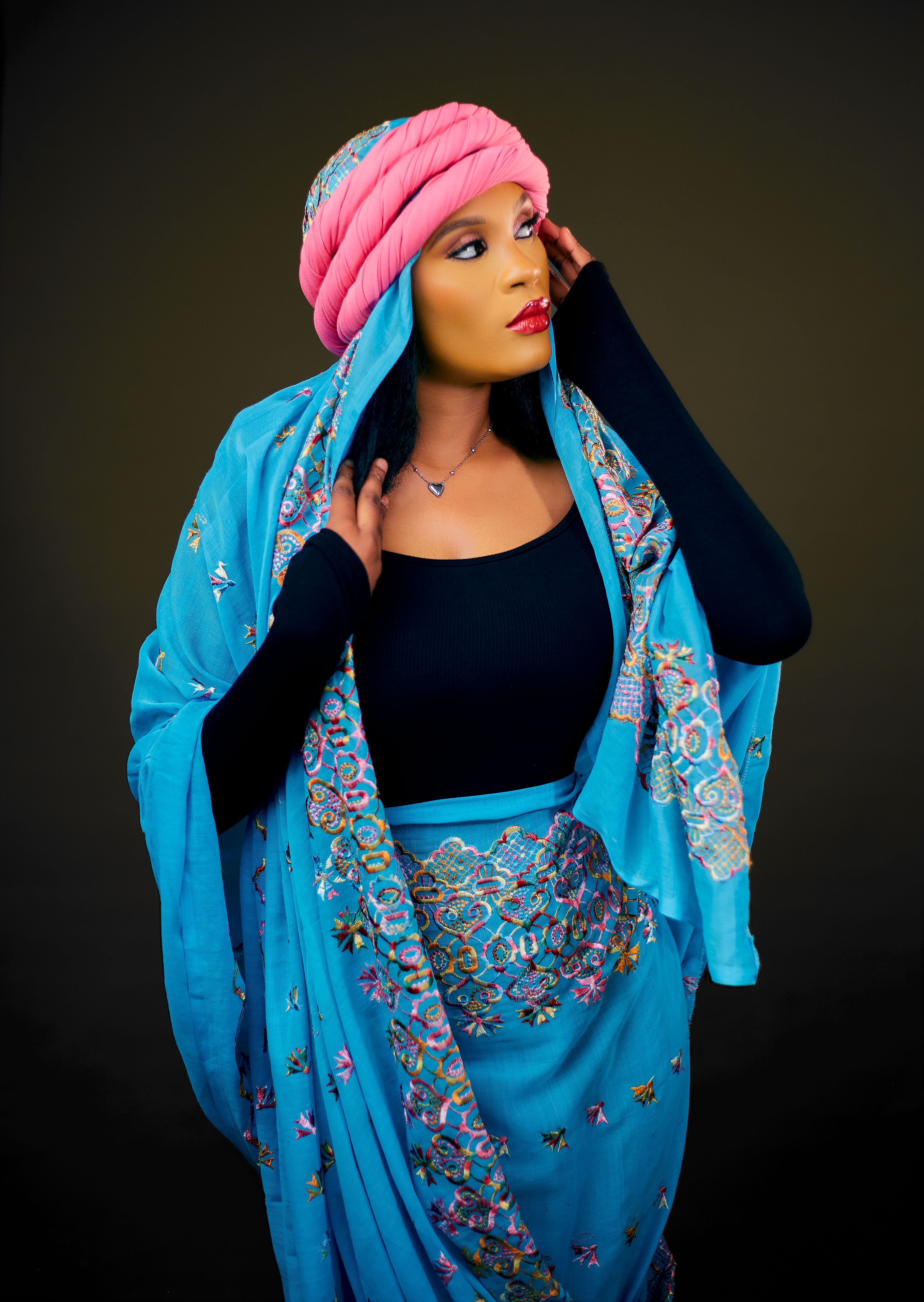 Portrait of a woman in vibrant traditional dress with a pink turban against a black backdrop.