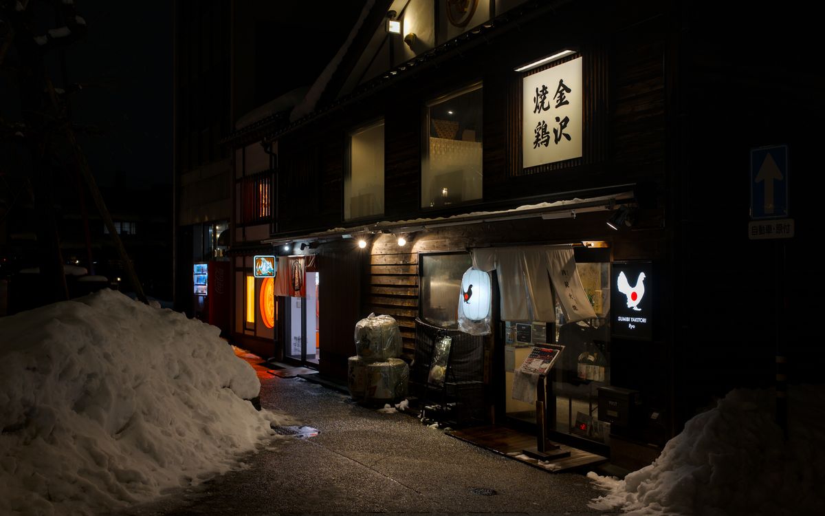 Lantern-lit izakaya fronts on a dark street in Kanazawa at night, with piles of snow along the road