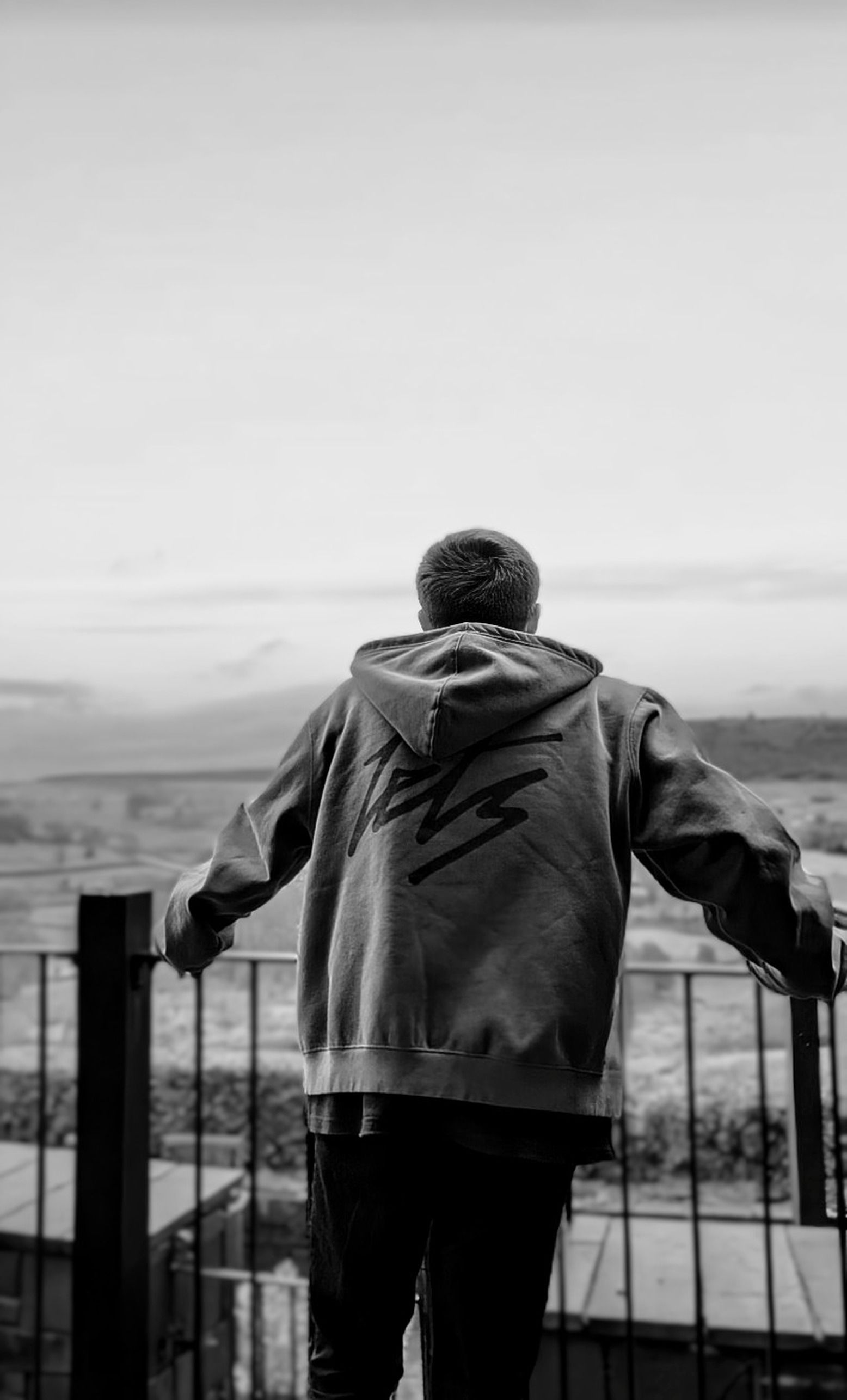 A person in a hoodie gazes over a scenic landscape from a balcony, evoking a reflective mood.