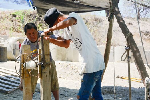 Two men working on a construction site, using a manual pulley under a tarp cover.