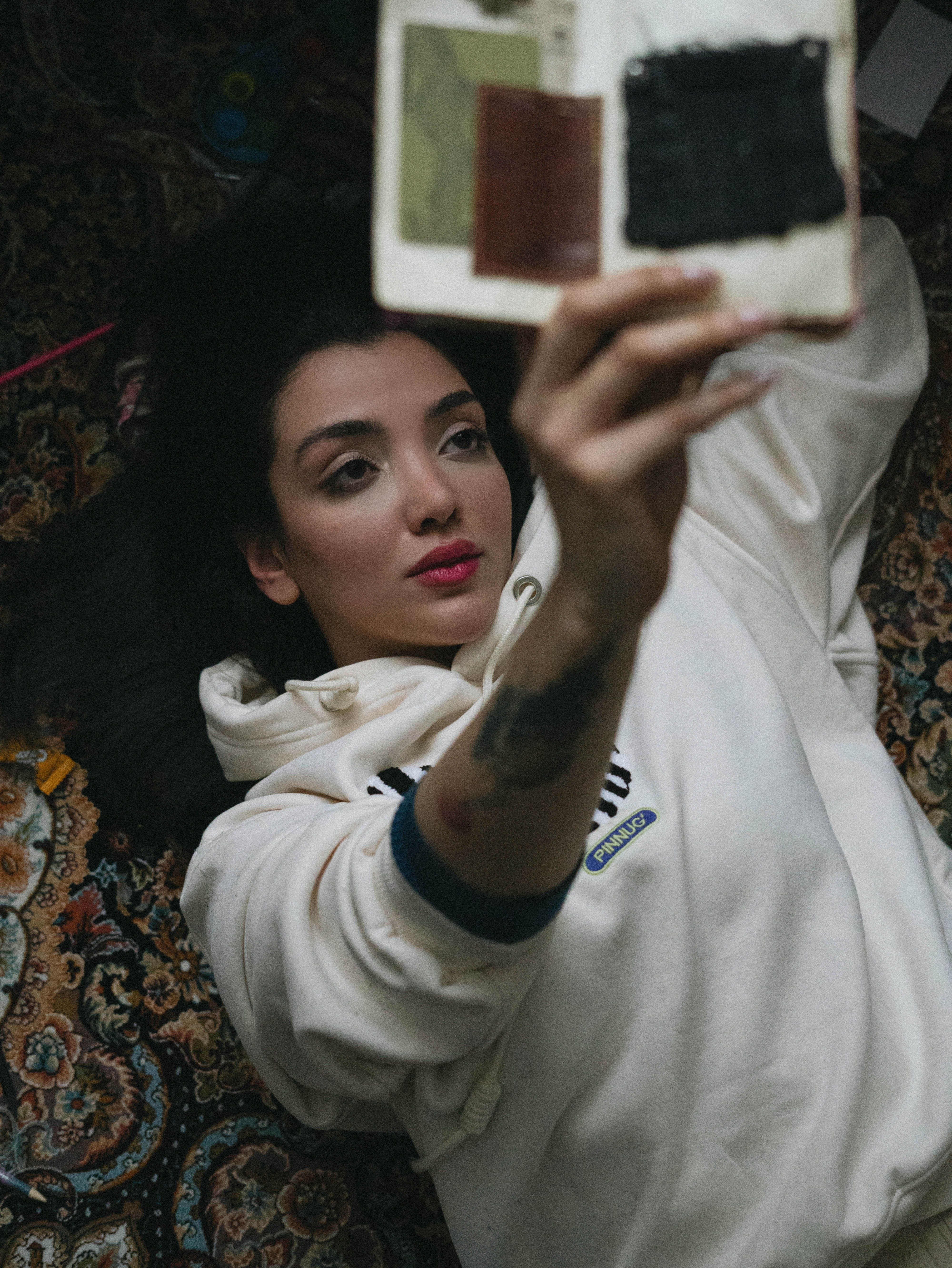 A young woman lying on a patterned rug, holding an art journal with paint samples indoors.