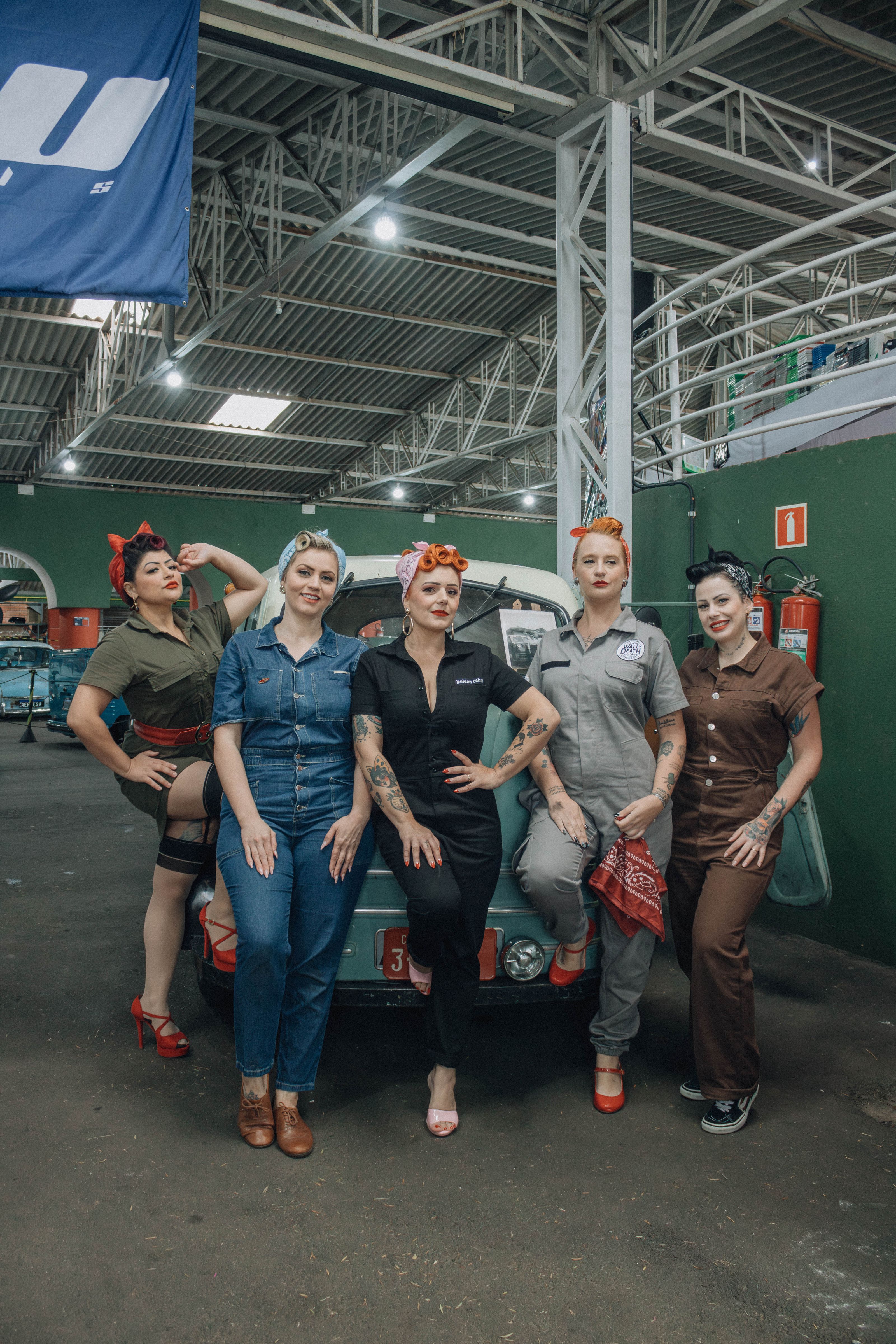 Cinco mulheres em trajes retrô posando com confiança em um ambiente de garagem.
