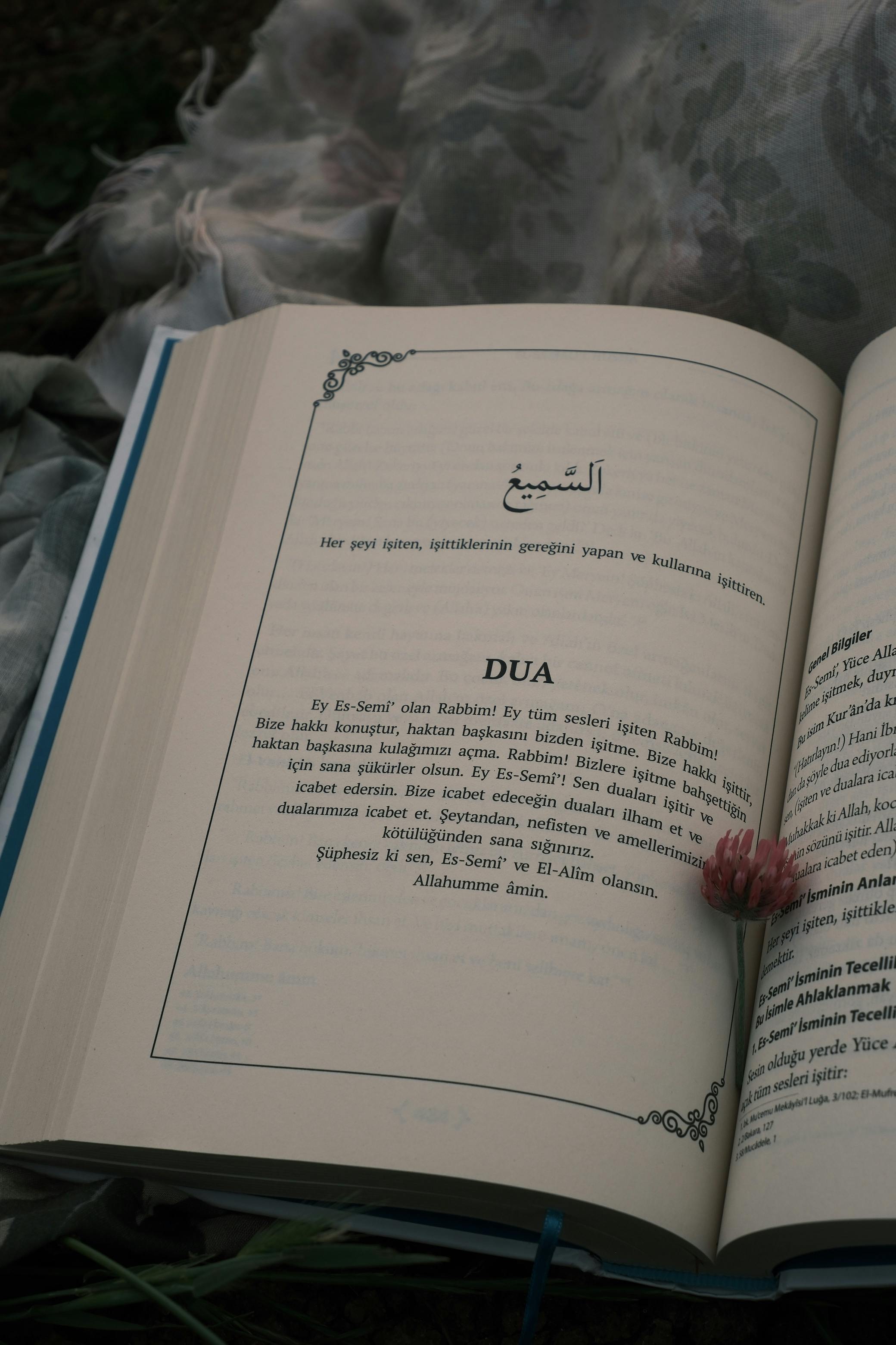 A book opened to a page with Arabic and Turkish text, adorned with a flower on a textile backdrop.