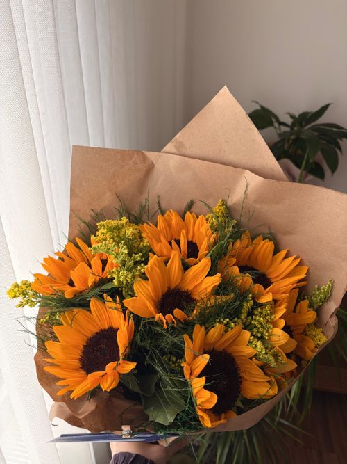 Free A vibrant bouquet of sunflowers and foliage wrapped in brown paper, placed by a sunlit window. Stock Photo