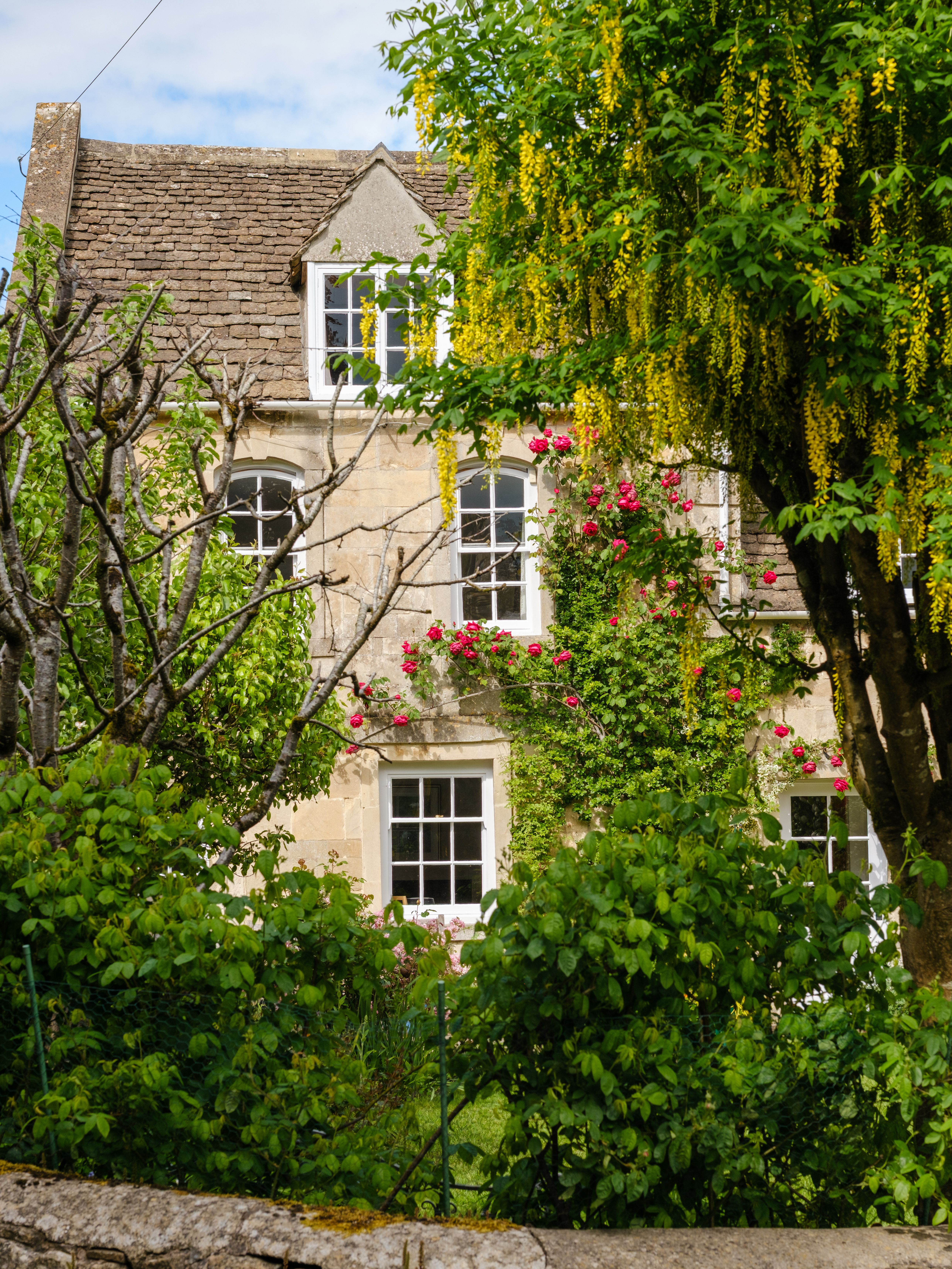 A picturesque English cottage surrounded by lush greenery and blossoming flowers in Tetbury, UK.
