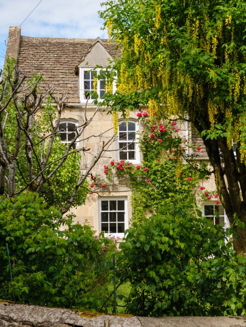A picturesque English cottage surrounded by lush greenery and blossoming flowers in Tetbury, UK.