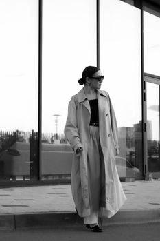 Stylish woman in trench coat and sunglasses, modern urban backdrop.
