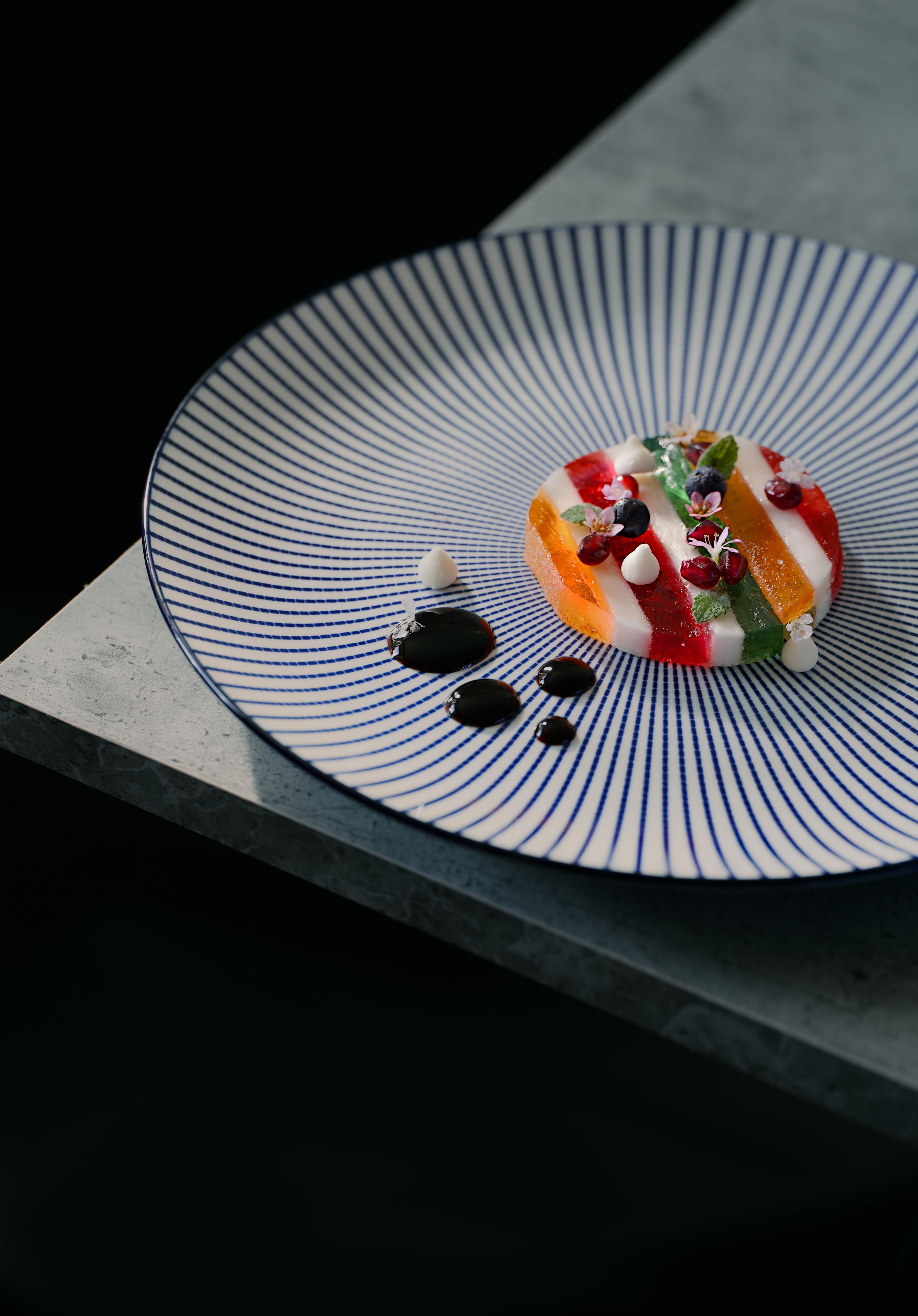 Dessert dressé sur une assiette à fines rayures bleues : fruits, meringues et points de sauce sombre