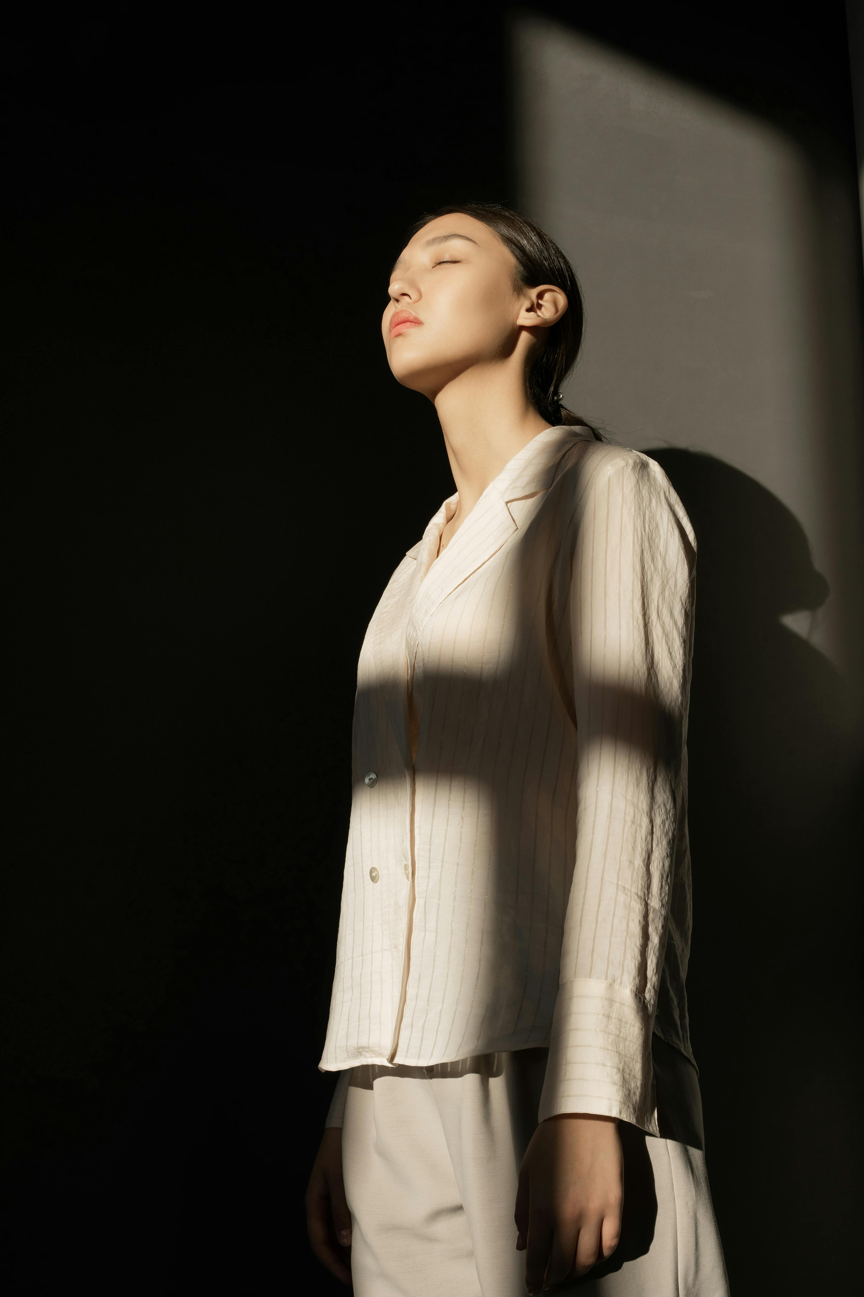 一位时尚女性闭眼倚墙而立，沐浴在阳光与阴影之中