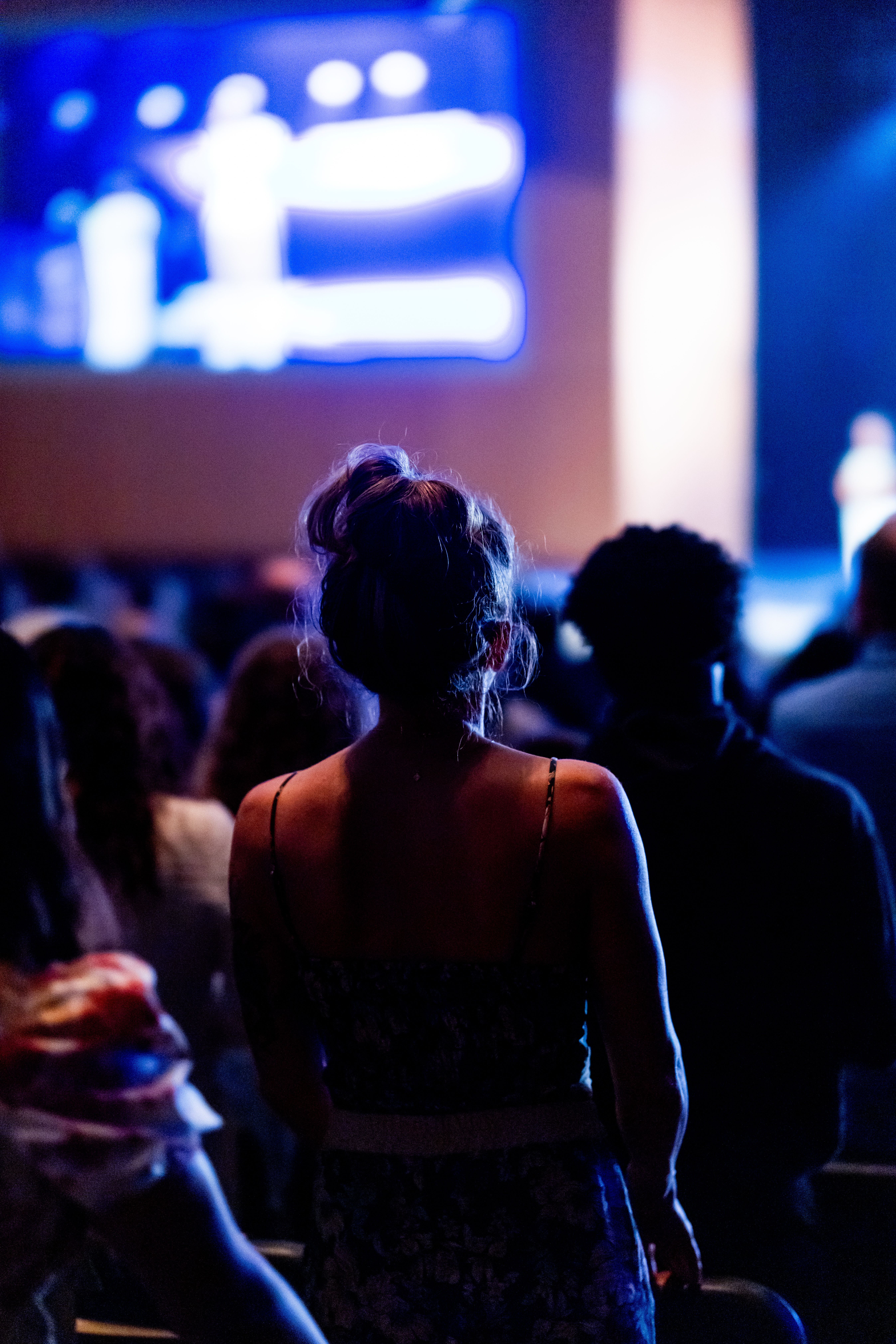 Back view of a diverse audience at a live event, focusing on a woman with her hair in a bun.