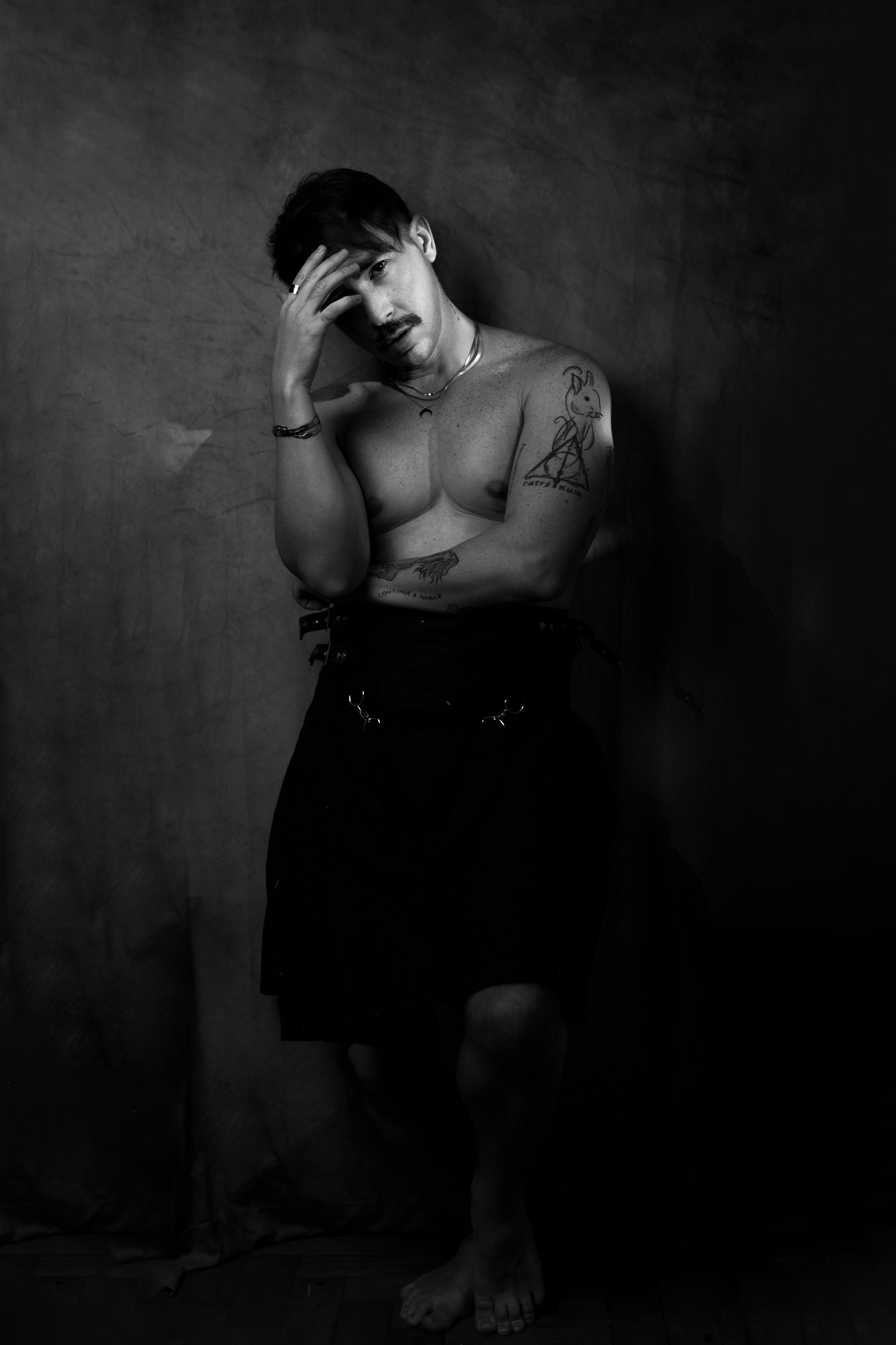 Moody black and white portrait of a shirtless tattooed man leaning against a wall, captured indoors.