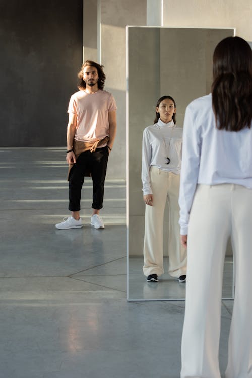 Woman in White Bluish Long Sleeve Shirt and White Pants Standing In Front of a Mirror