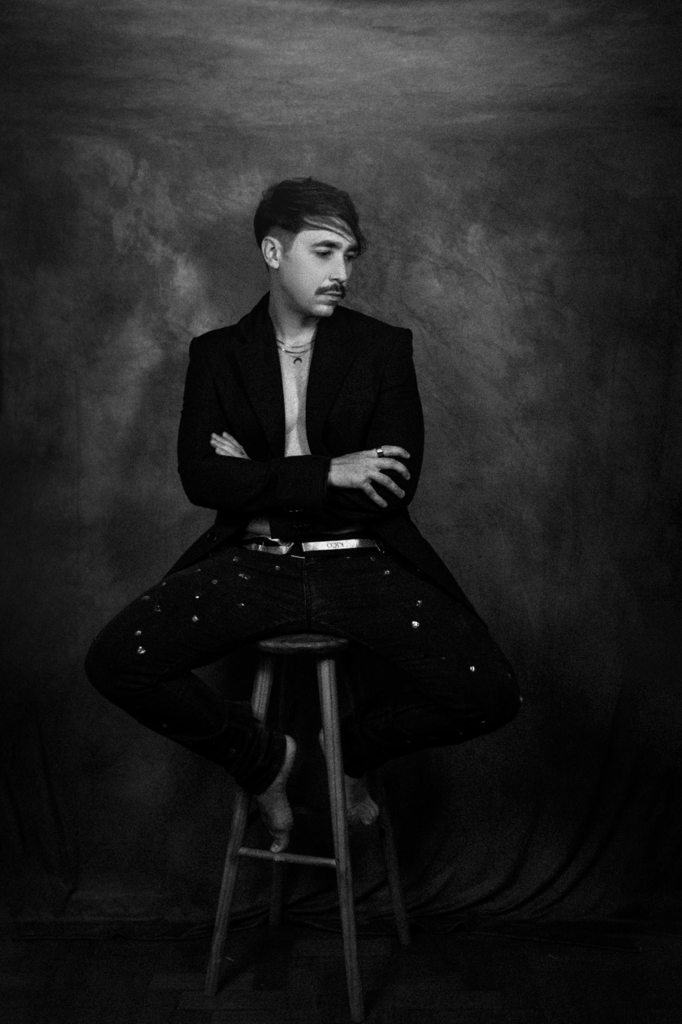 Elegant black and white portrait of a man seated on a stool with arms crossed in a studio setting.