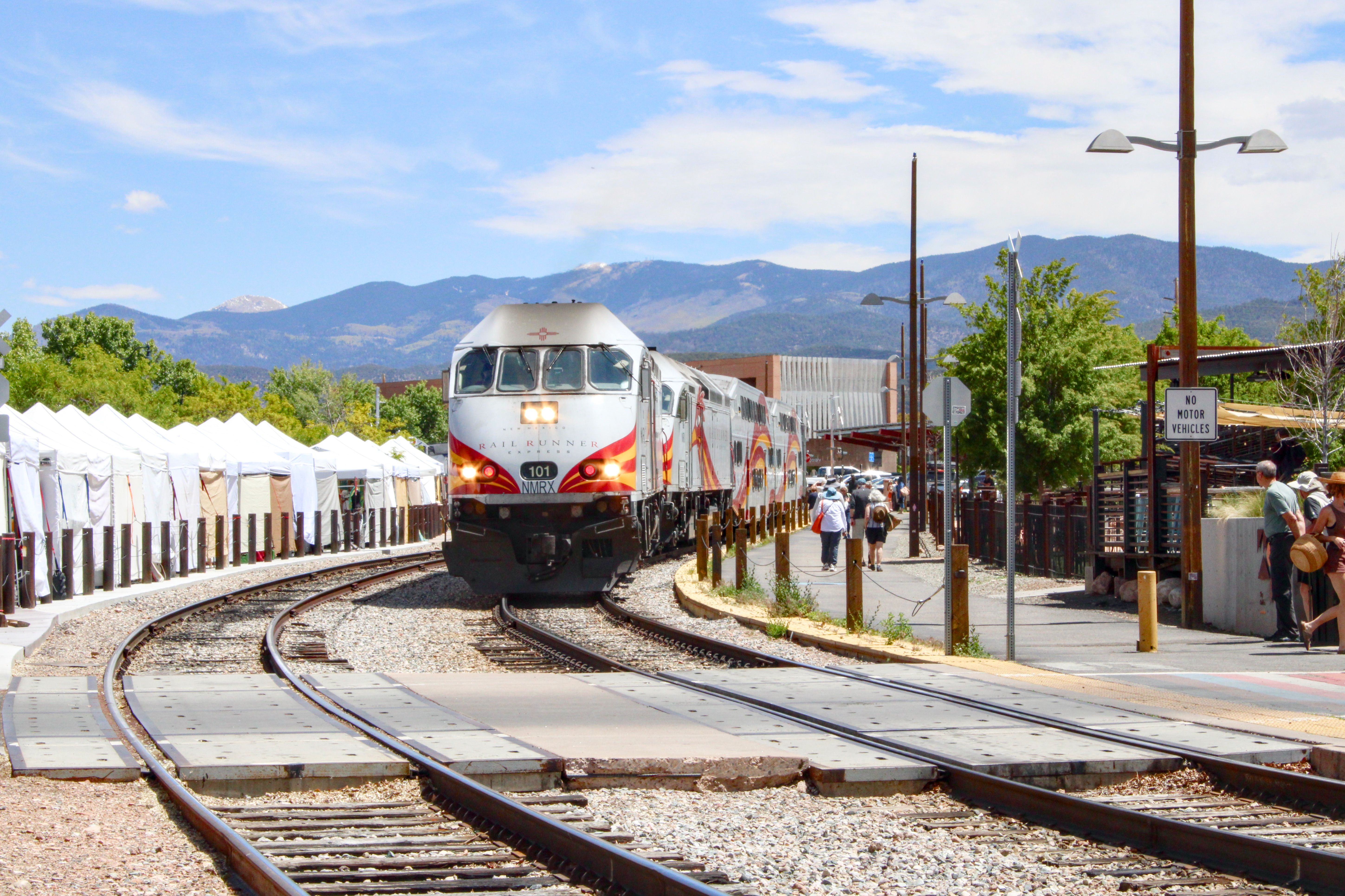A passenger train approaches a station with mountains in the background on a sunny day.