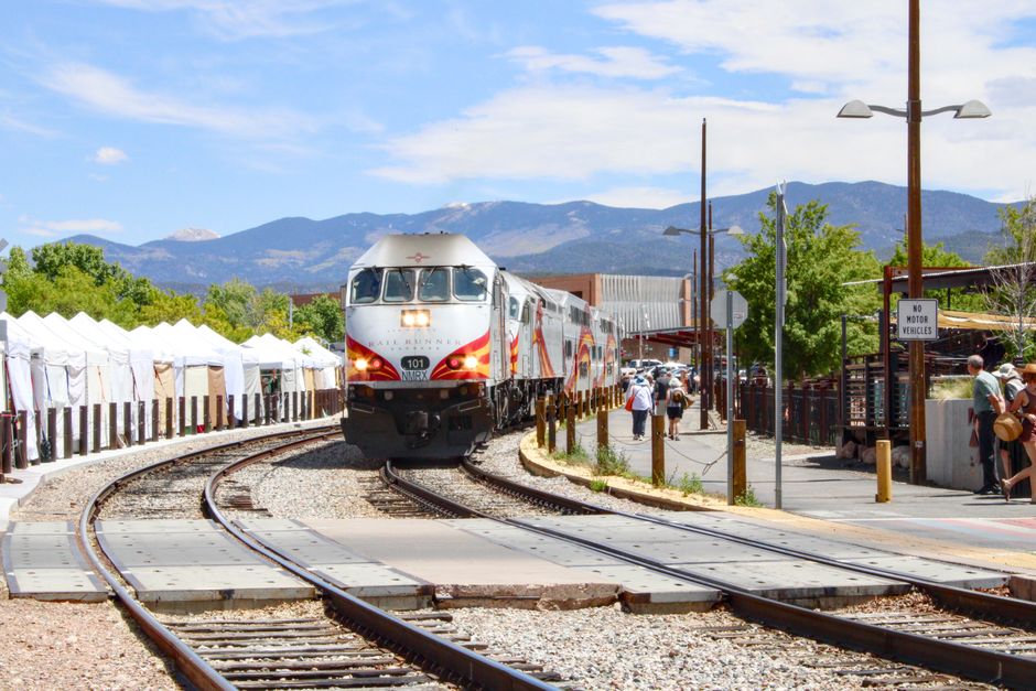 A passenger train approaches a station with mountains in the background on a sunny day.