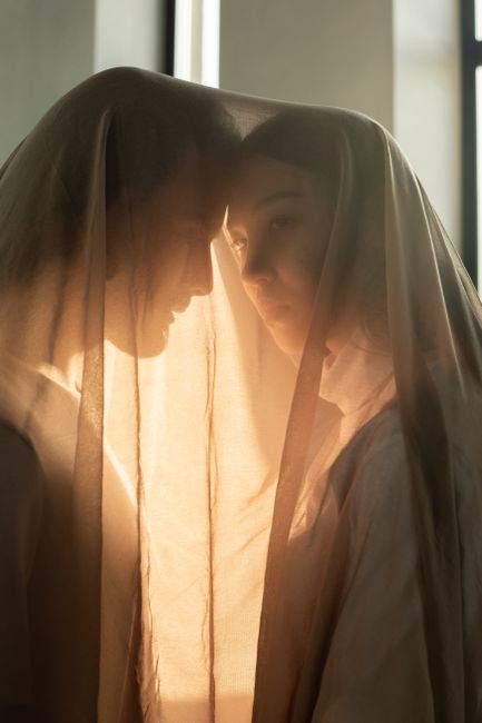 A couple intimately embracing under a sheer veil with soft, warm lighting indoors.