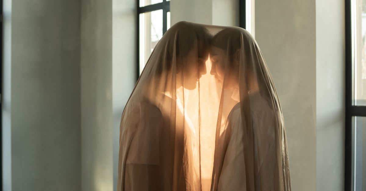 Two people in a romantic embrace under a sheer veil indoors, bathed in warm light.