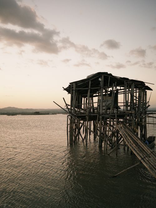 Free A rustic bamboo hut on stilts stands in serene water under a warm sunset sky. Stock Photo