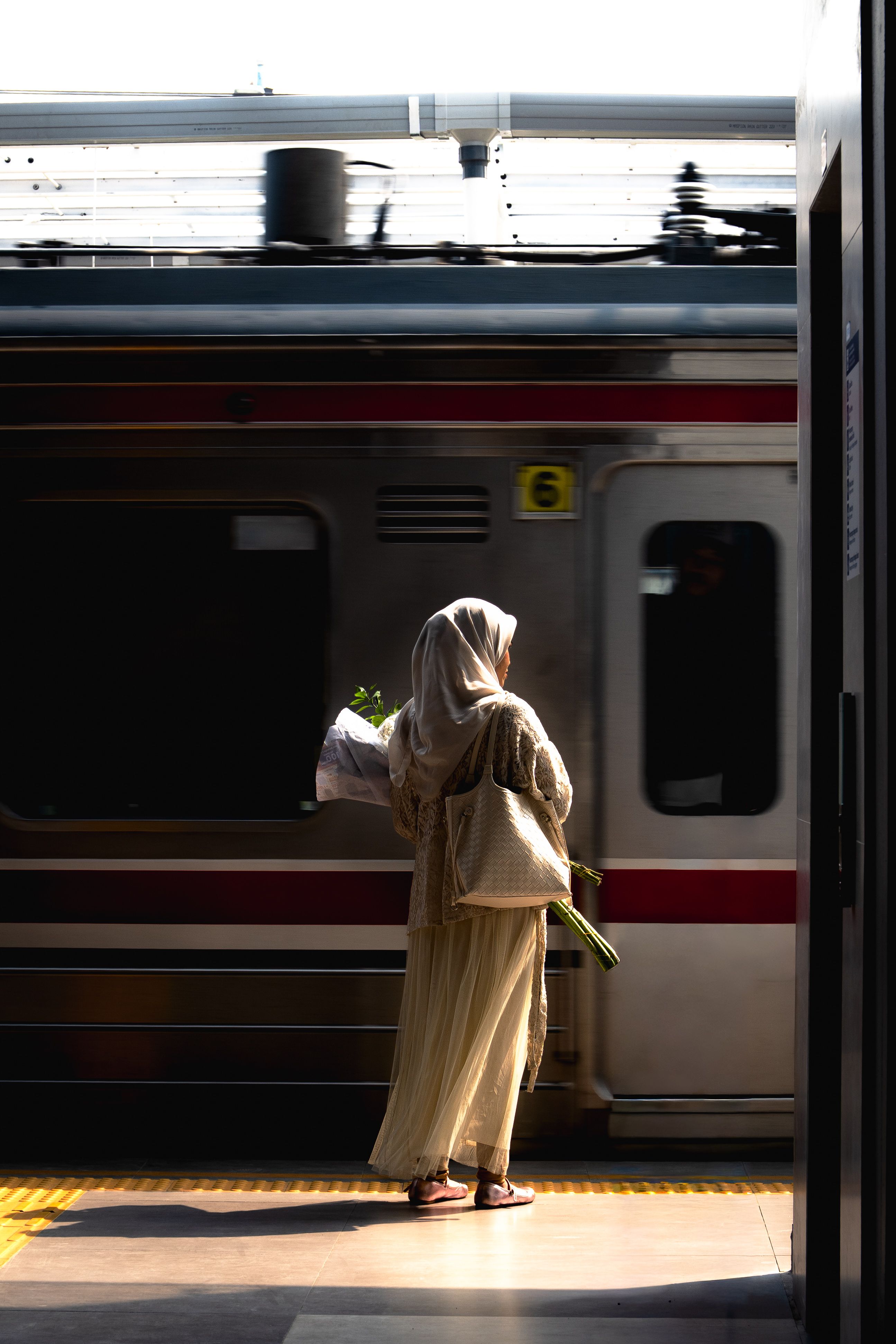A woman in traditional attire waits at a sunlit train platform, holding flowers.