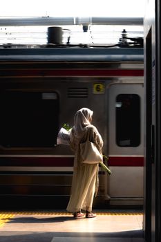 A woman in traditional attire waits at a sunlit train platform, holding flowers.