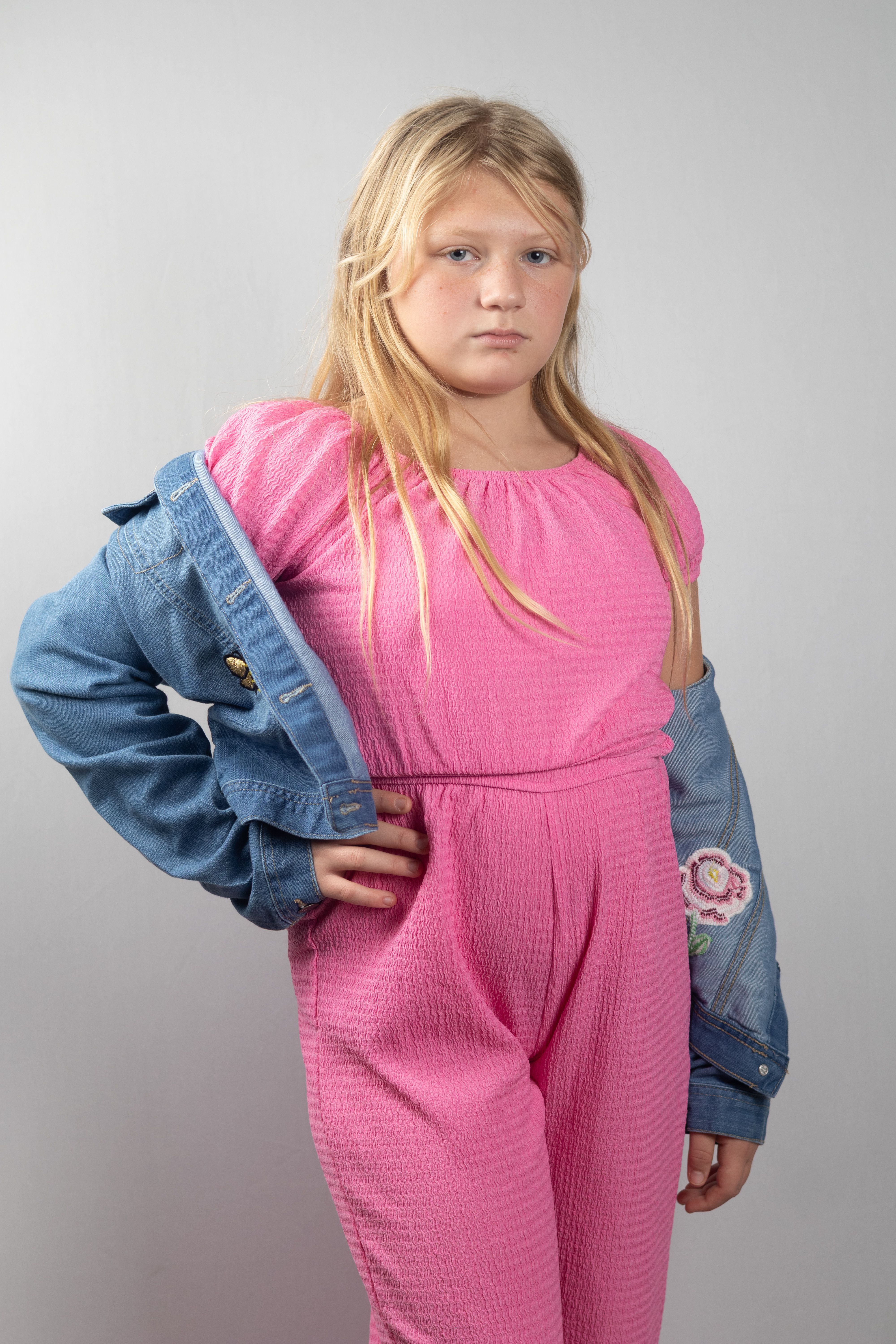 Young girl posing confidently in a pink jumpsuit and denim jacket against a neutral background.
