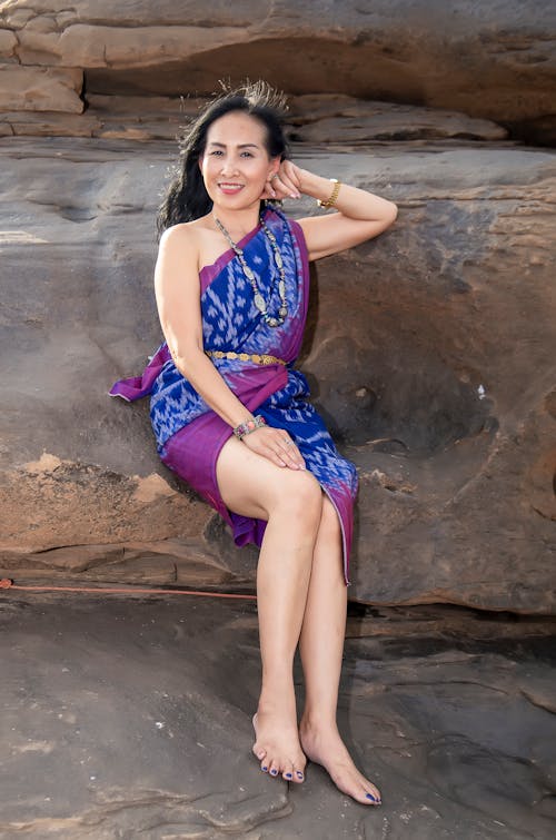 Photo of Smiling Woman in Blue and Purple Floral Sleeveless Dress Sitting on Brown Rock
