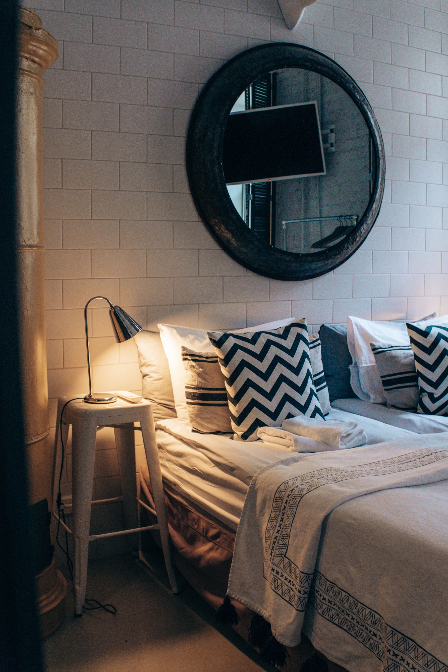 Bed layered with black and white patterned textiles, solid pillows, bedside lamp and mirror