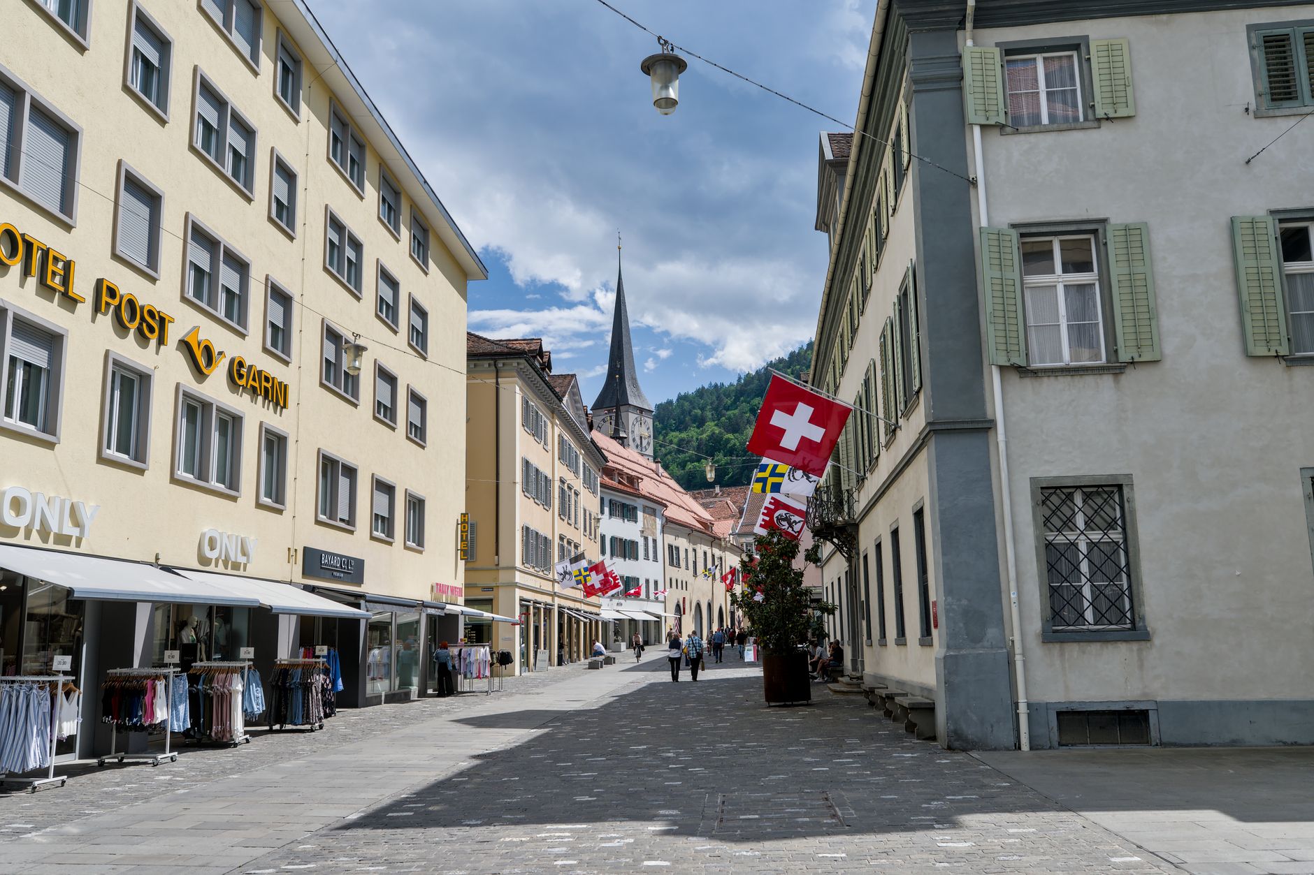 A picturesque street in Grisons, Switzerland featuring a church spire and Swiss flags under a bright sky.