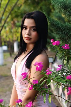 A woman in a pink dress poses gracefully among blooming pink flowers in an outdoor setting.