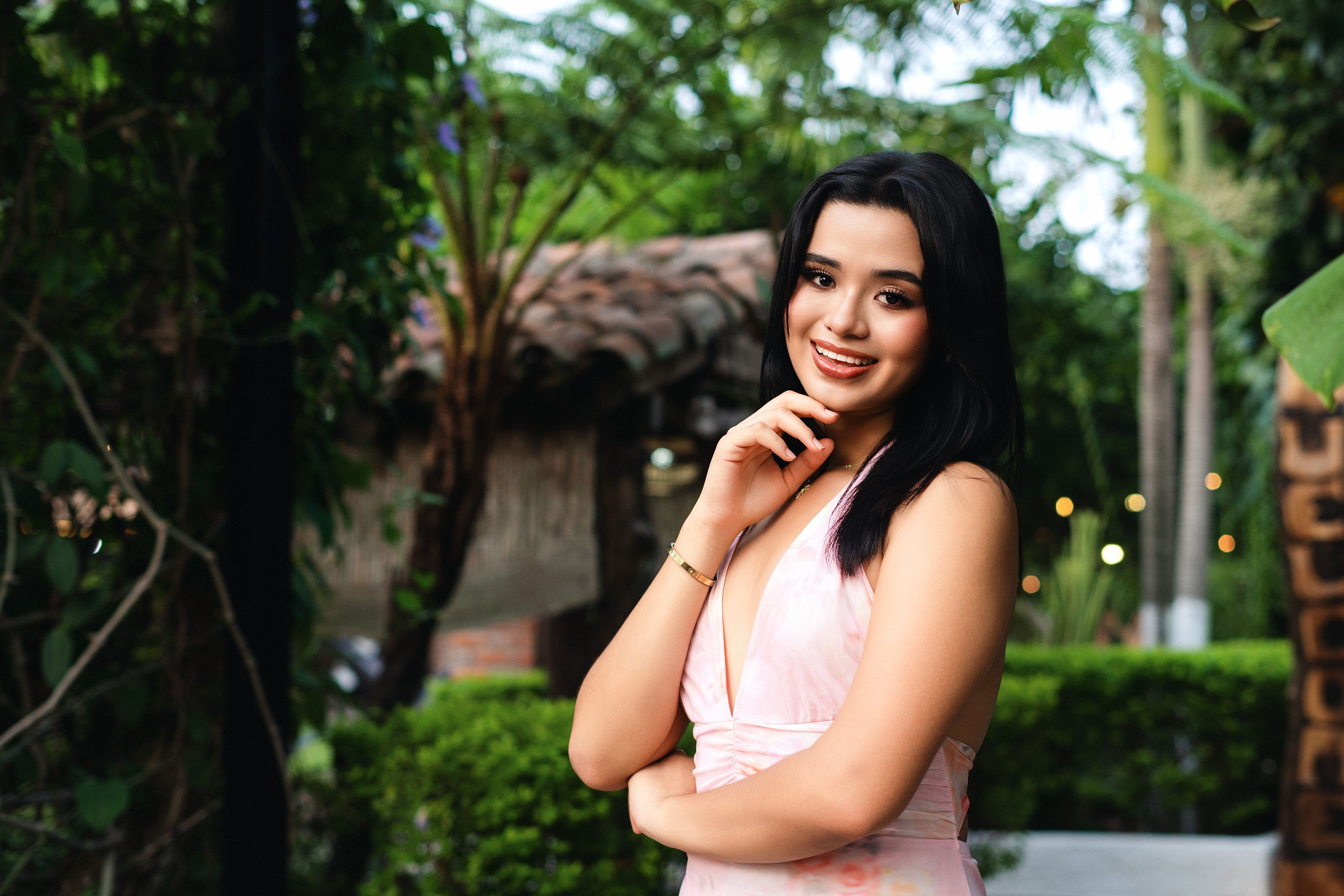A young woman smiles confidently outdoors, surrounded by lush greenery and rustic architecture.