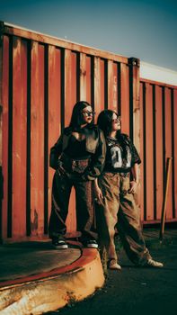 Two women pose in trendy urban outfits against a rust-colored shipping container.