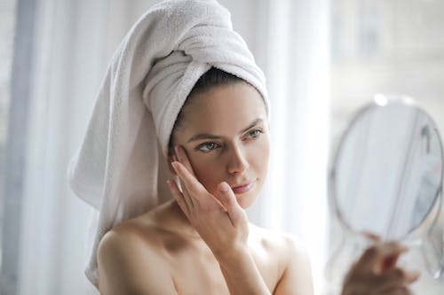 Young female with bare shoulders and towel on head holding small mirror and checking skin with it in bathroom