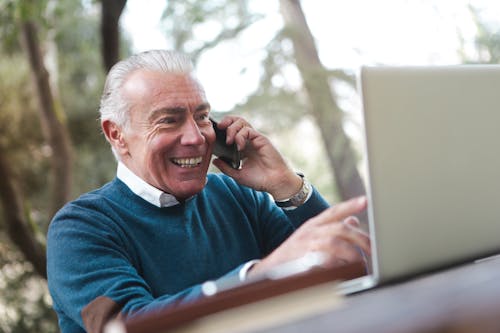 微笑的老人穿著藍色毛衣，坐在桌旁，一邊使用筆記本電腦在電話上交談的選擇性焦點照片