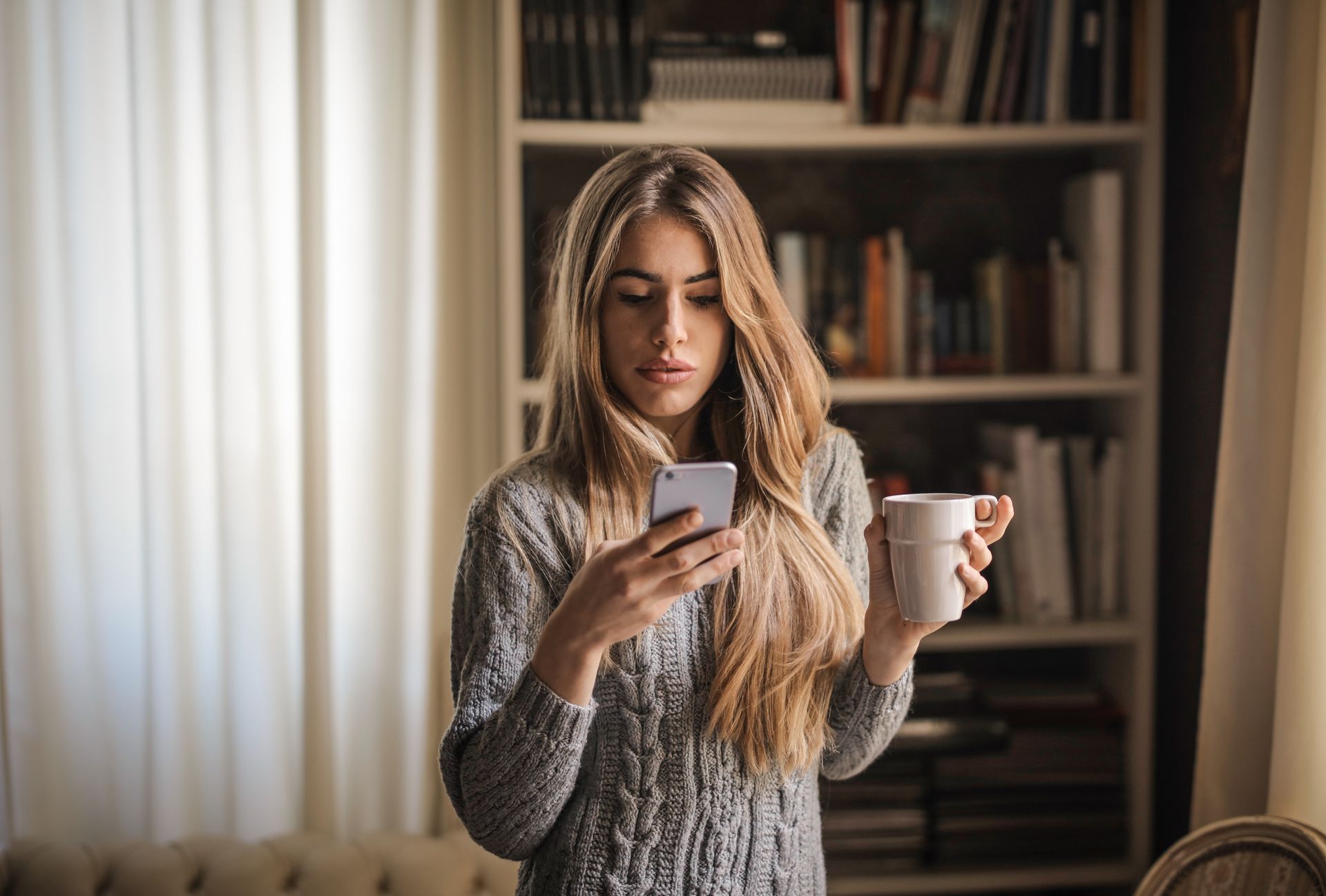 Young woman in a knit sweater texting on a smartphone while sipping coffee at home.