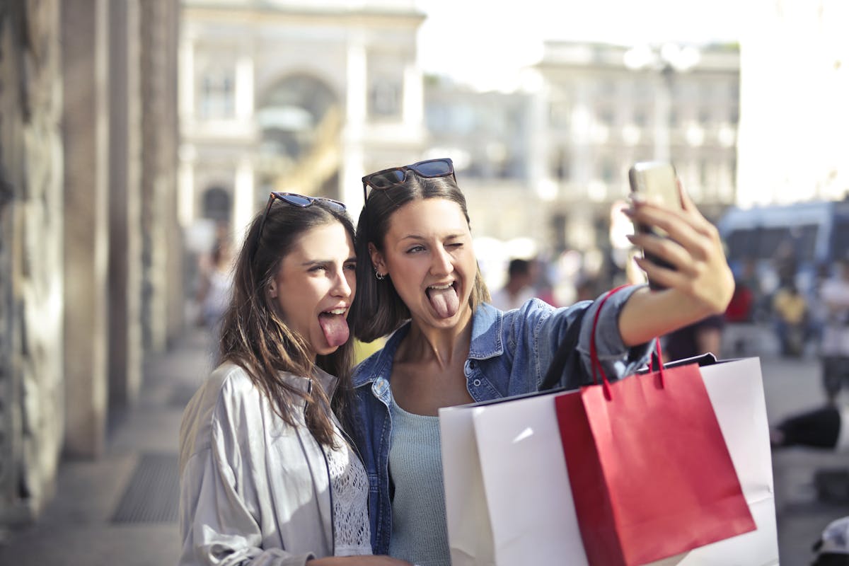 Cheerful young women with shopping bags taking selfie on street
