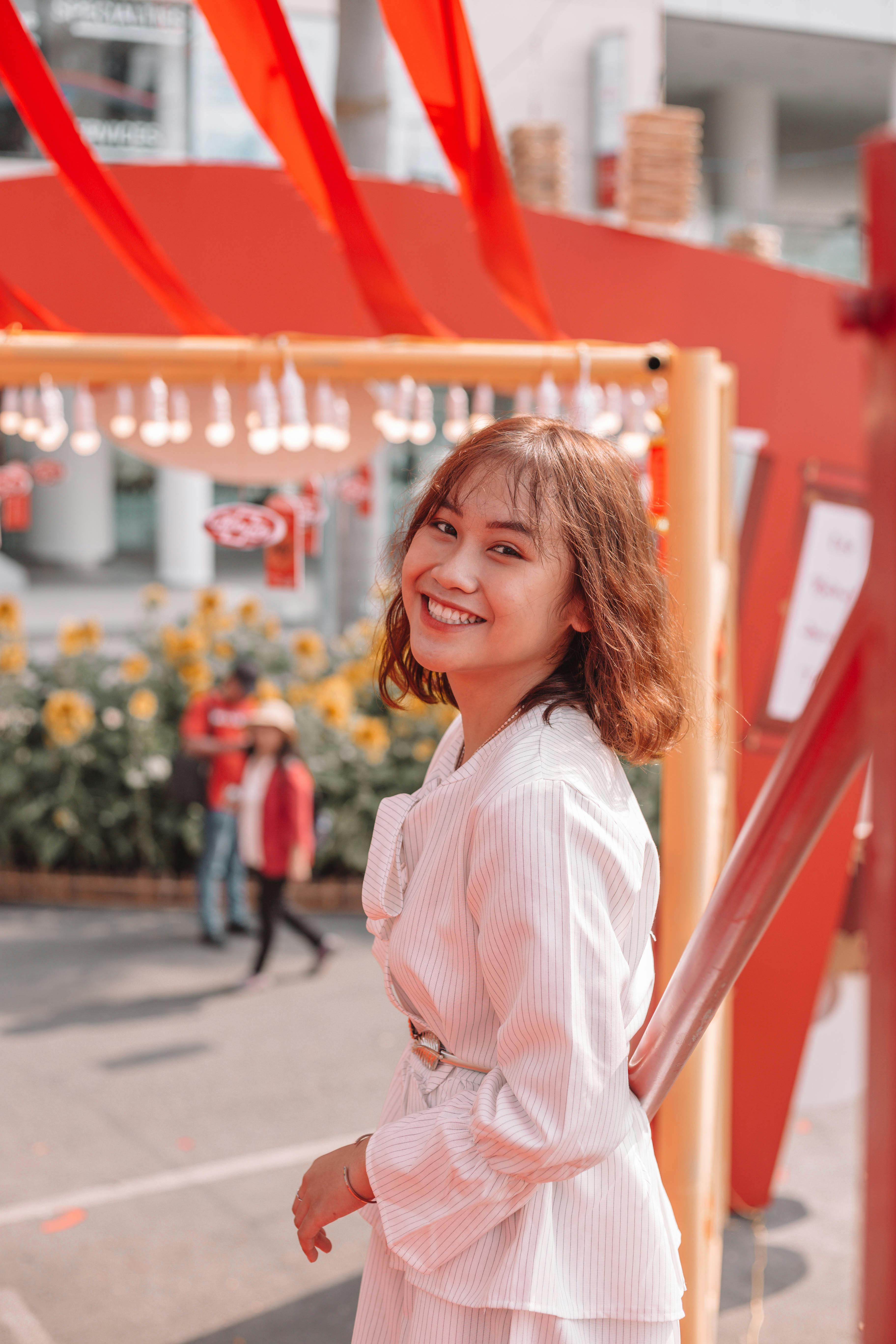 在色彩缤纷的户外节日场景中，一位身穿白衣的女子微笑着出现，身上带有红色装饰