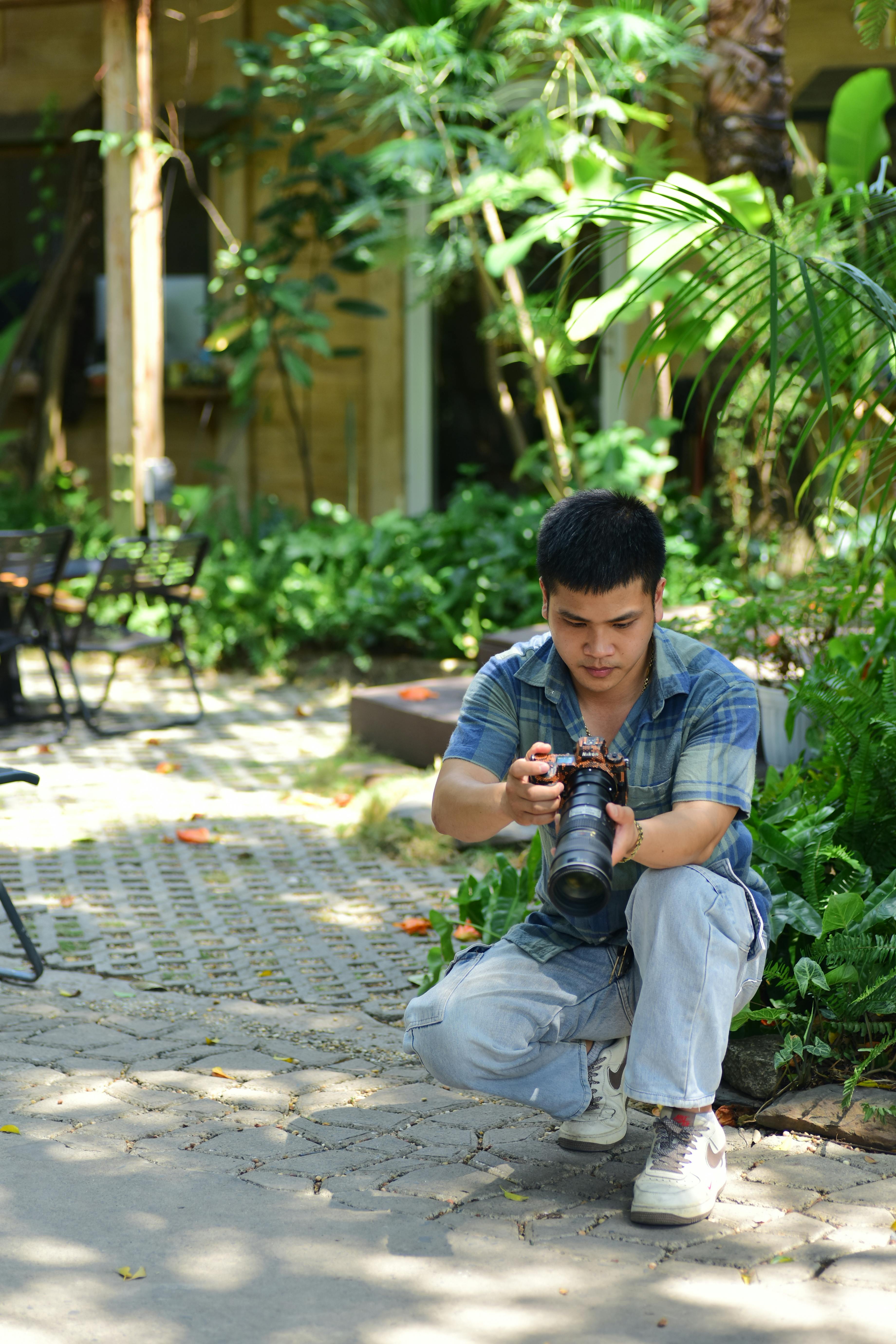 Young adult photographer crouching outdoors with camera in a lush garden setting, capturing nature.
