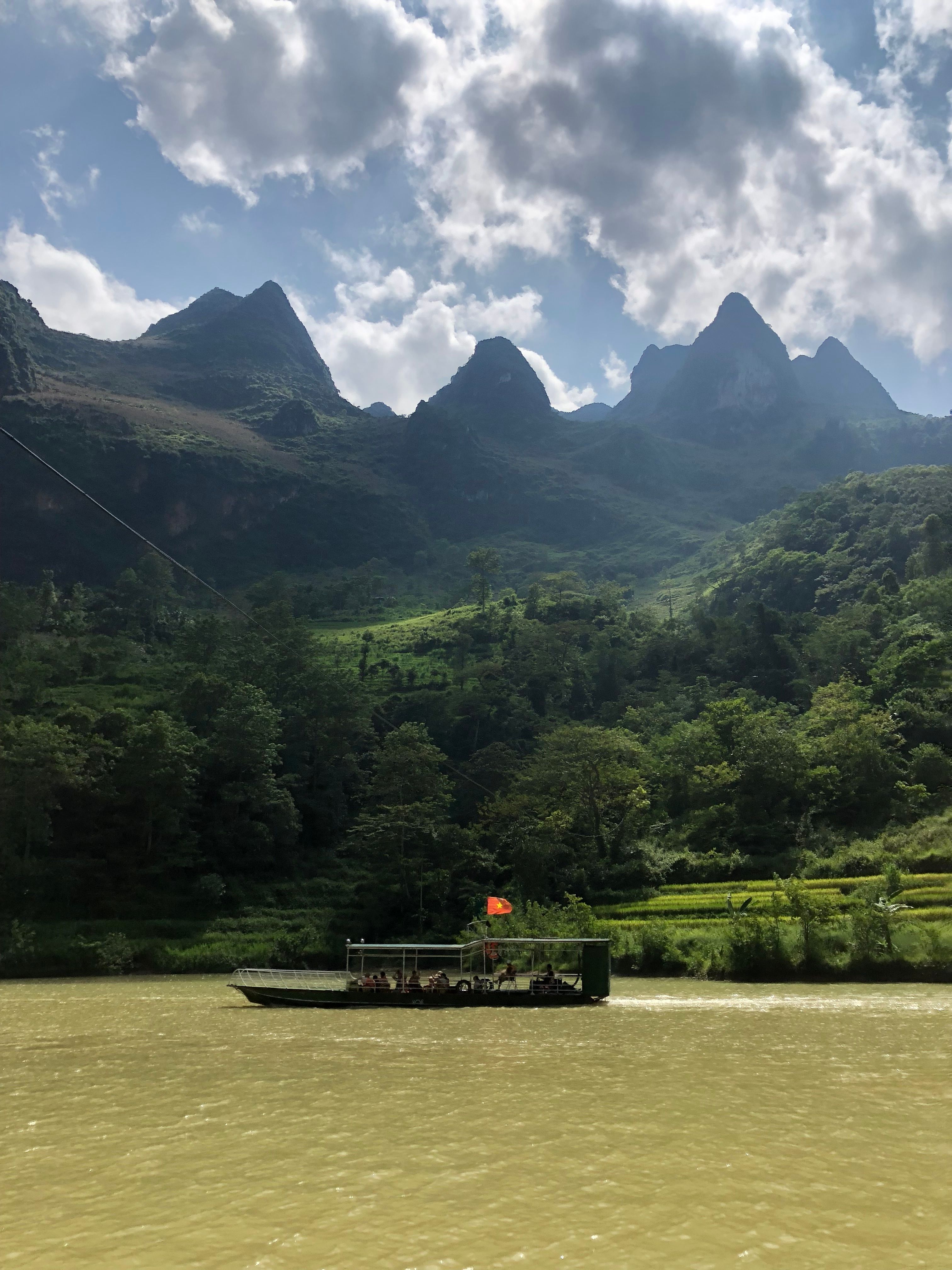 Ferry crossing a serene river with Vietnamese mountains in the background under a partly cloudy sky.