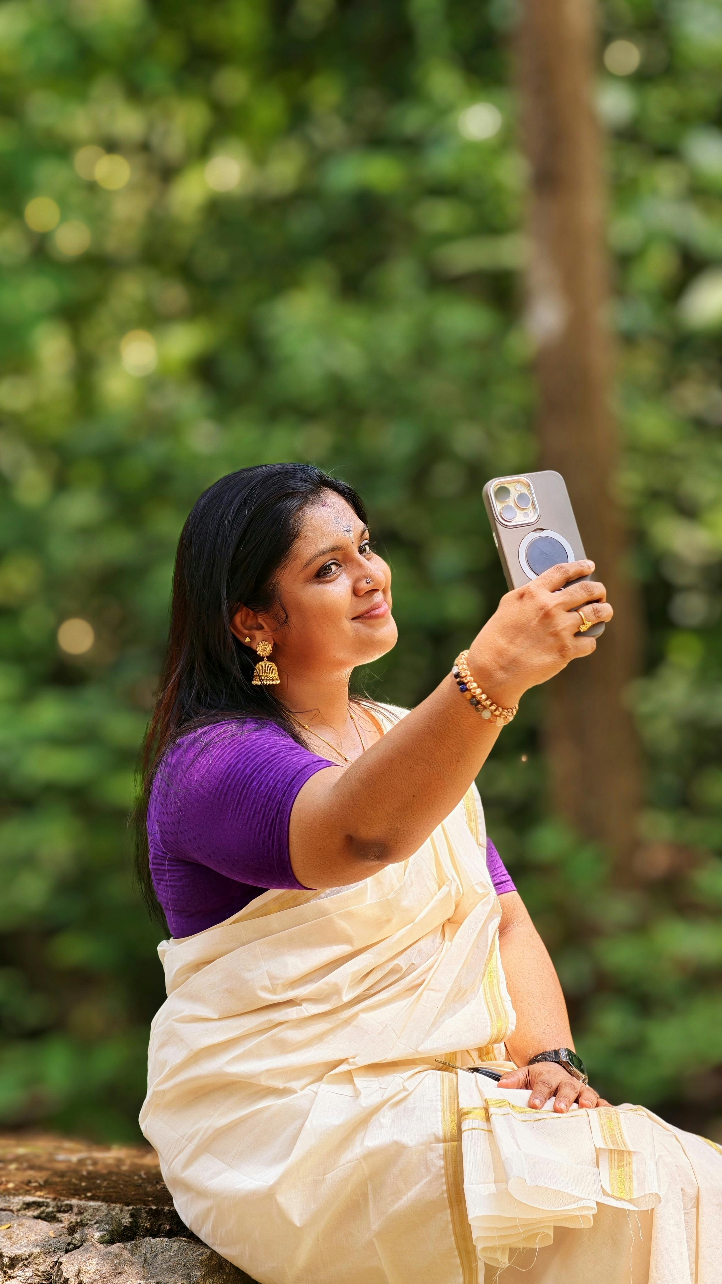 Traditional South Indian woman in a white saree and purple blouse takes a selfie outdoors.