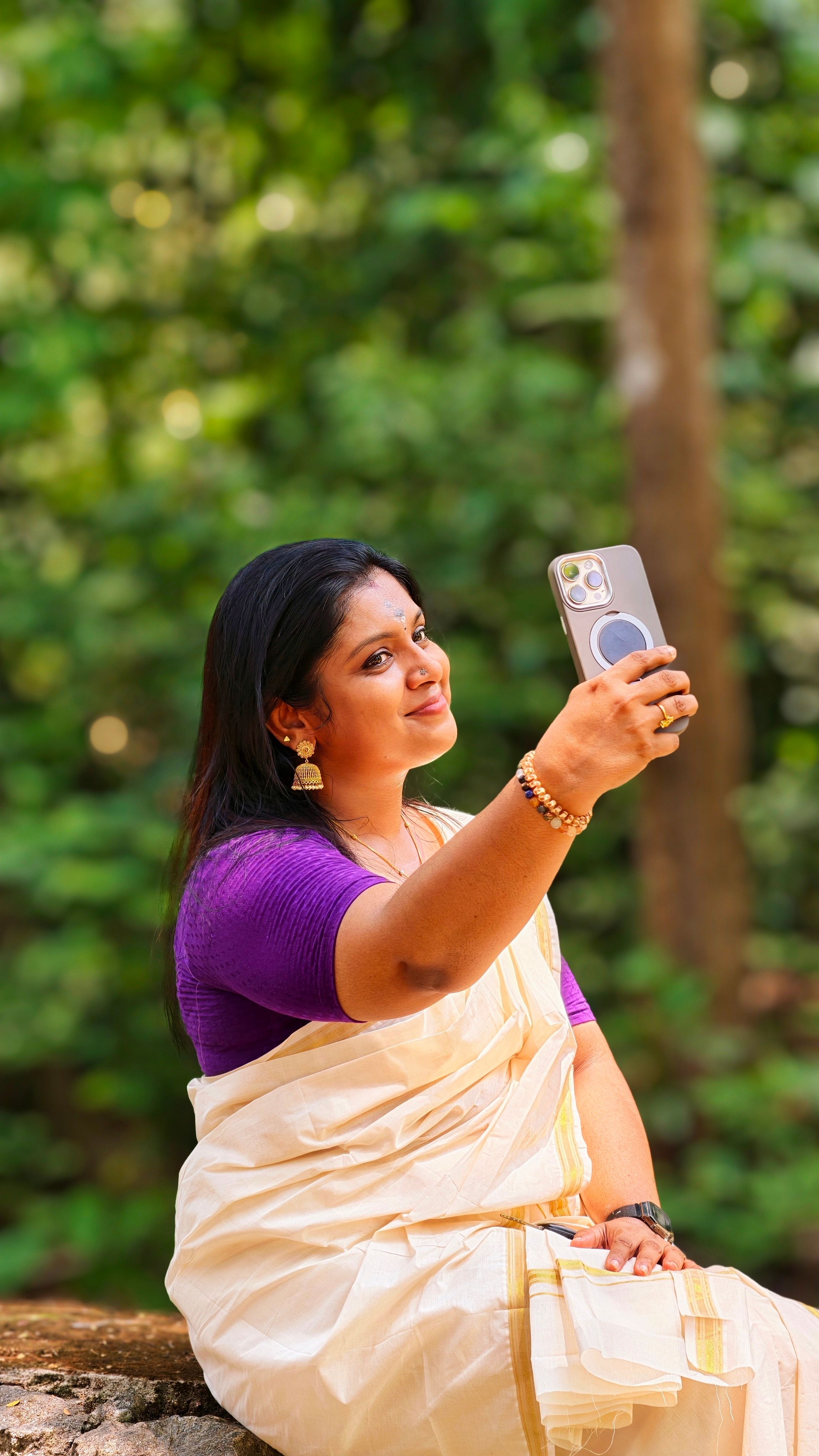 South Asian woman in traditional attire sitting outdoors, taking a selfie with lush greenery background.
