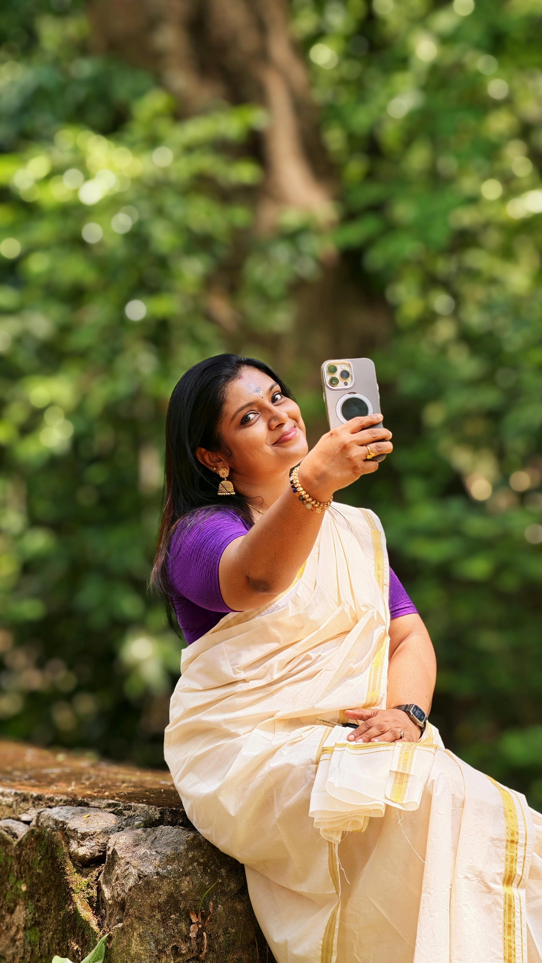 A woman in a traditional Kerala saree takes a selfie outdoors, blending tradition with technology.