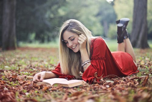 Улыбающаяся женщина в красном платье, лежа на коричневых сухих листьях на земле, читает книгу
