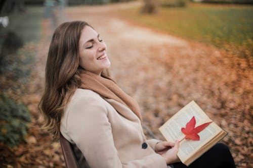 Selective Focus Photo of Laughing Woman in Brown Coat Holding a Book While Sitting in a Park Bench