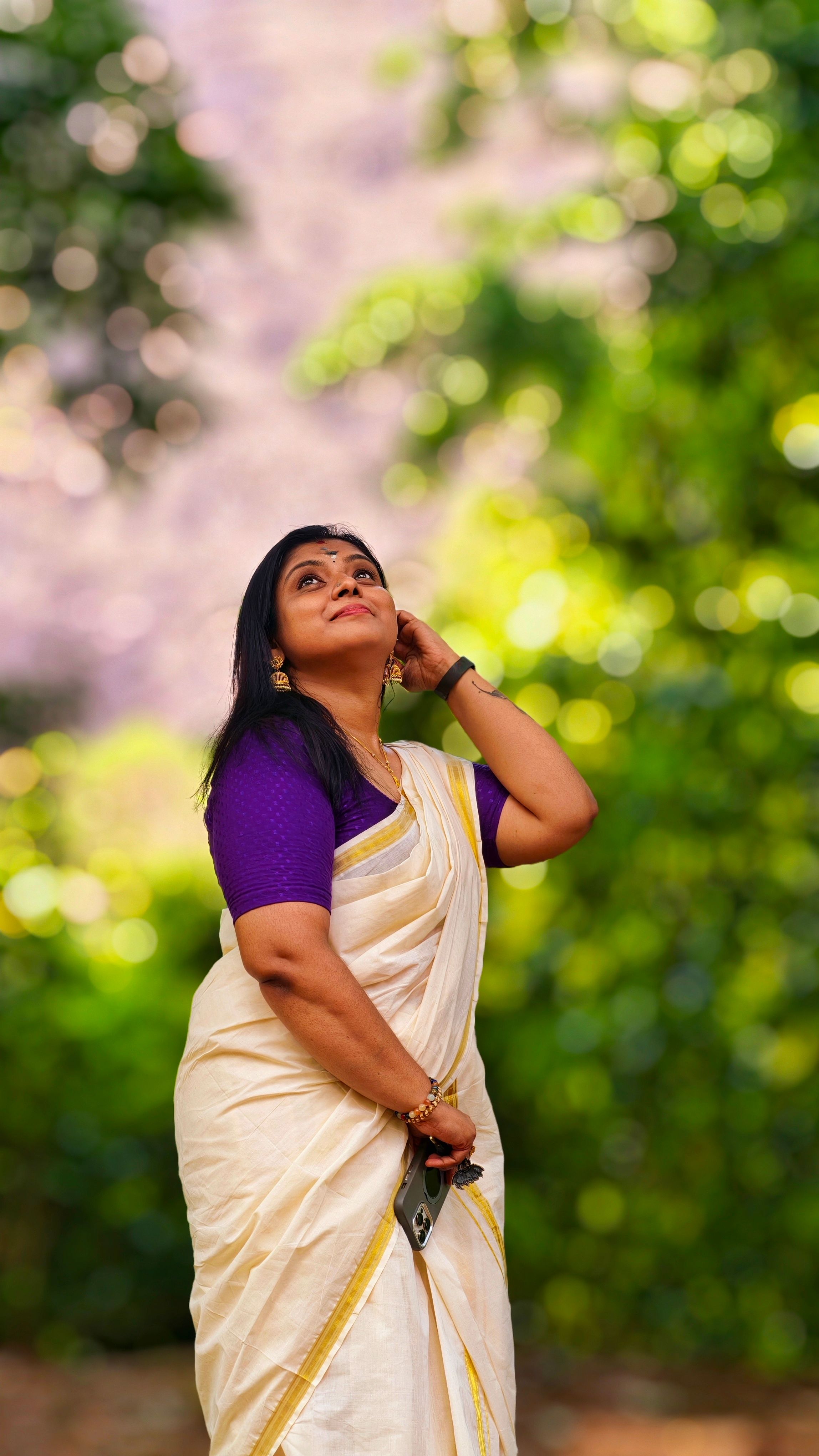 A graceful woman wearing a traditional Kerala saree and jewelry, posed elegantly in an outdoor setting.
