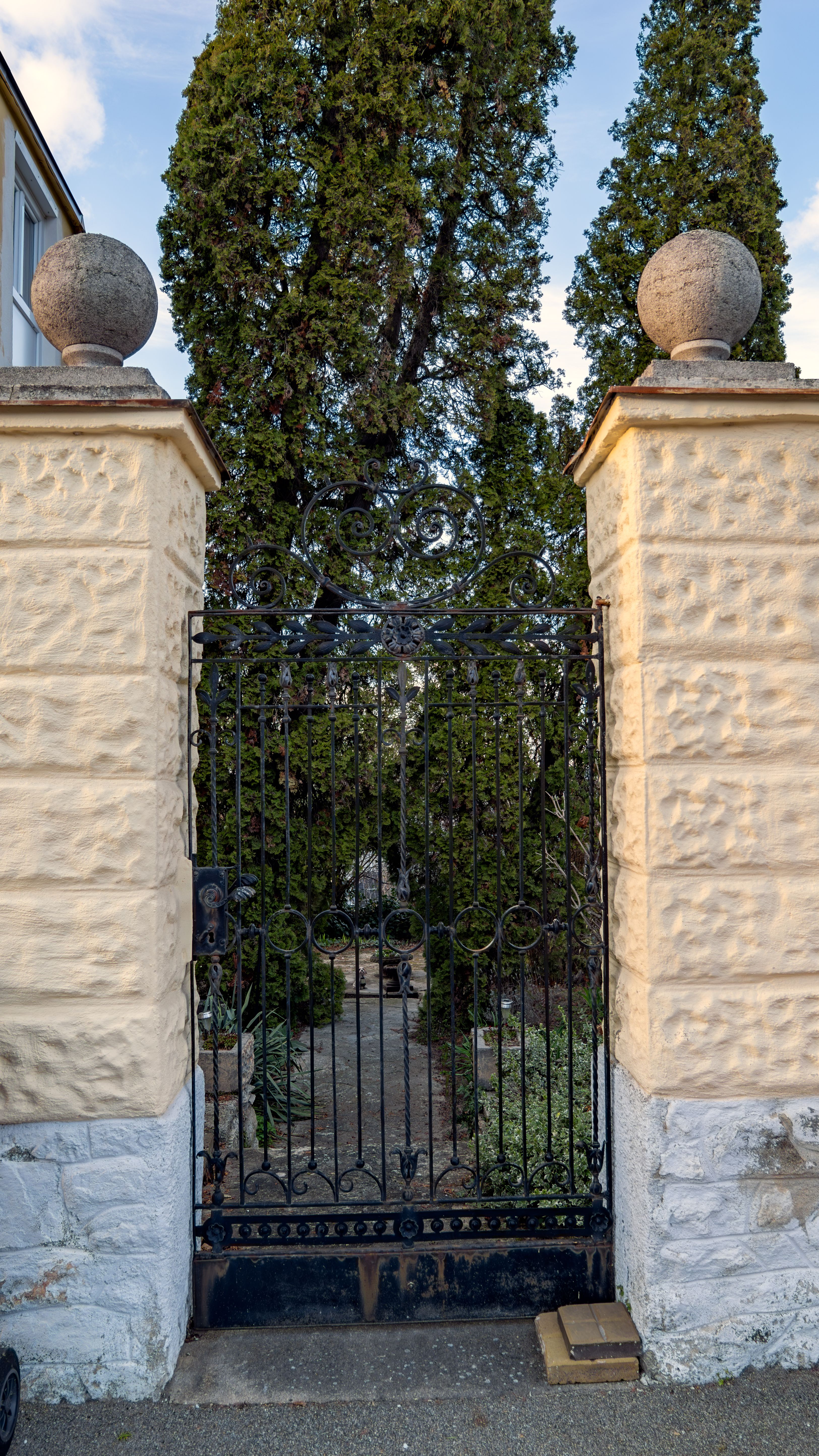 Portillon de jardin en fer forgé surmonté d’une volute, entre deux piliers de pierre