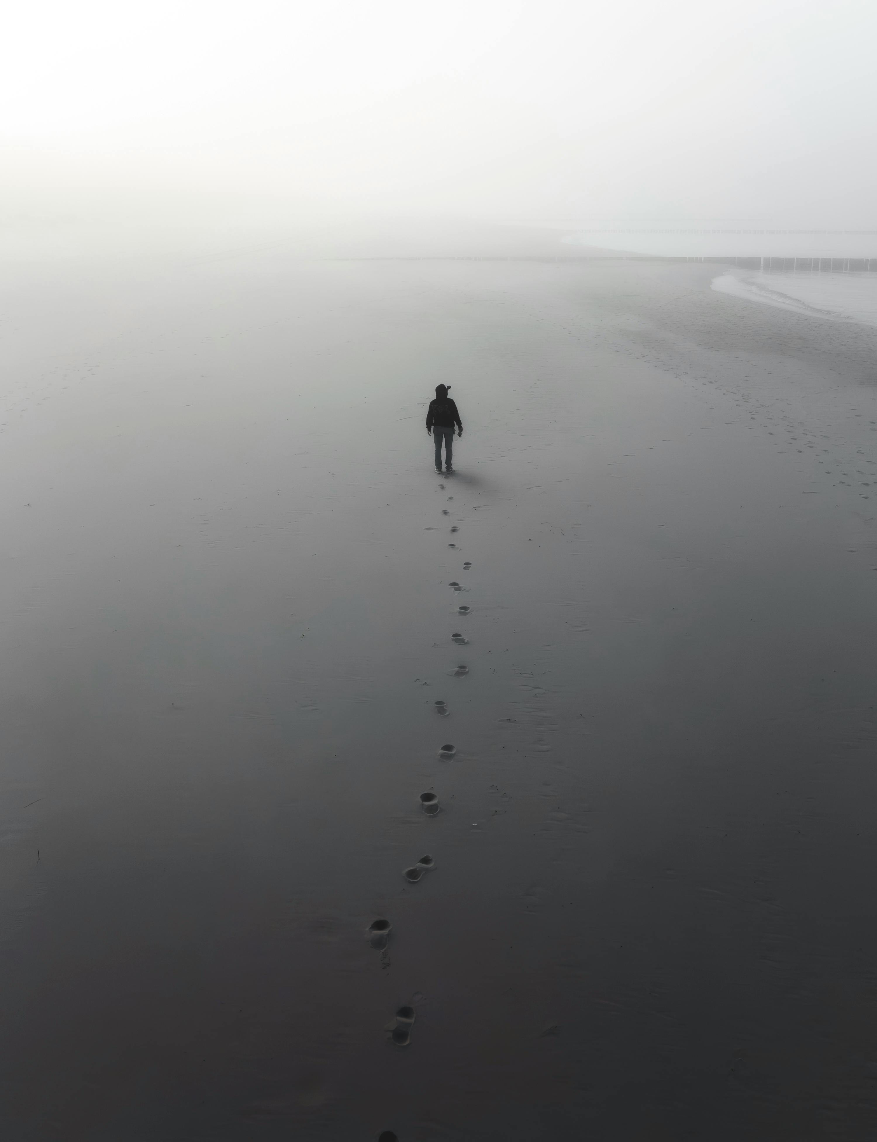 A lone figure walks along a misty beach, leaving footprints behind in the dense fog.